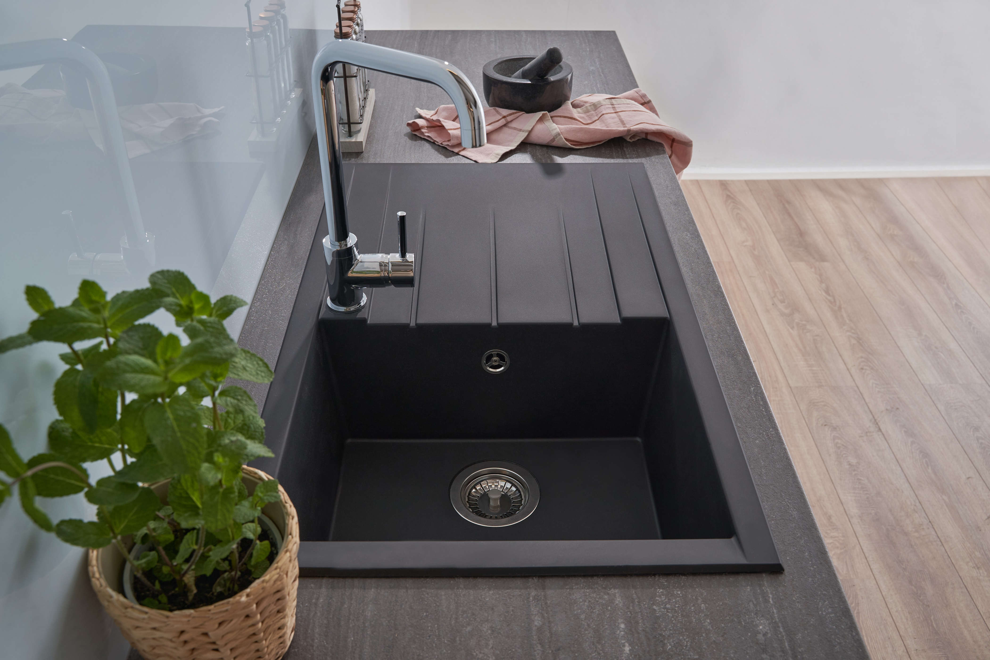 Schwarzes Küchenbecken mit Wasserhahn, Pflanze davor, Mörser und Handtuch rechts, graue Arbeitsplatte.
