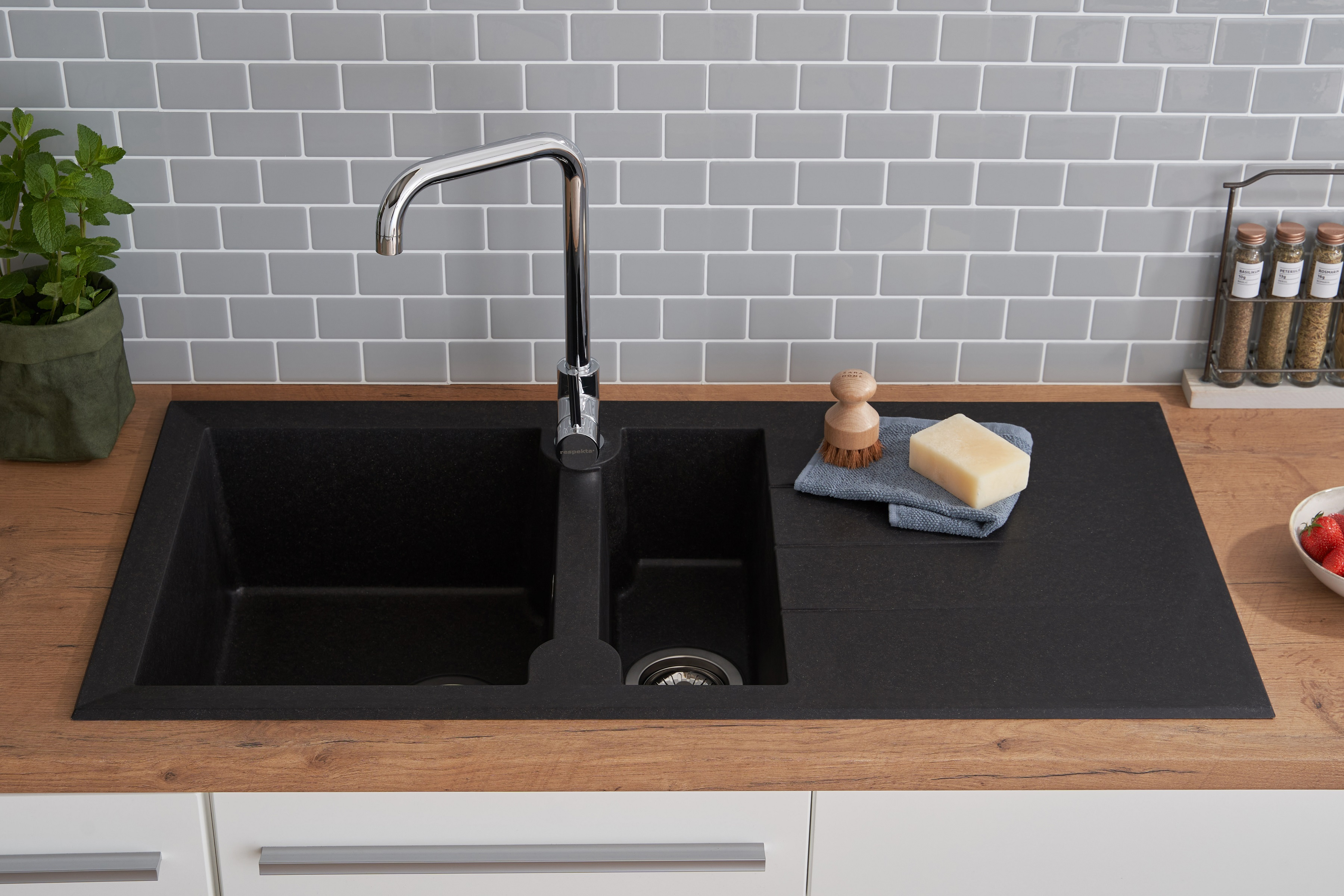 Schwarzes Waschbecken mit Wasserhahn. Auf der Theke befinden sich Seife, Bürste und Gewürze.
