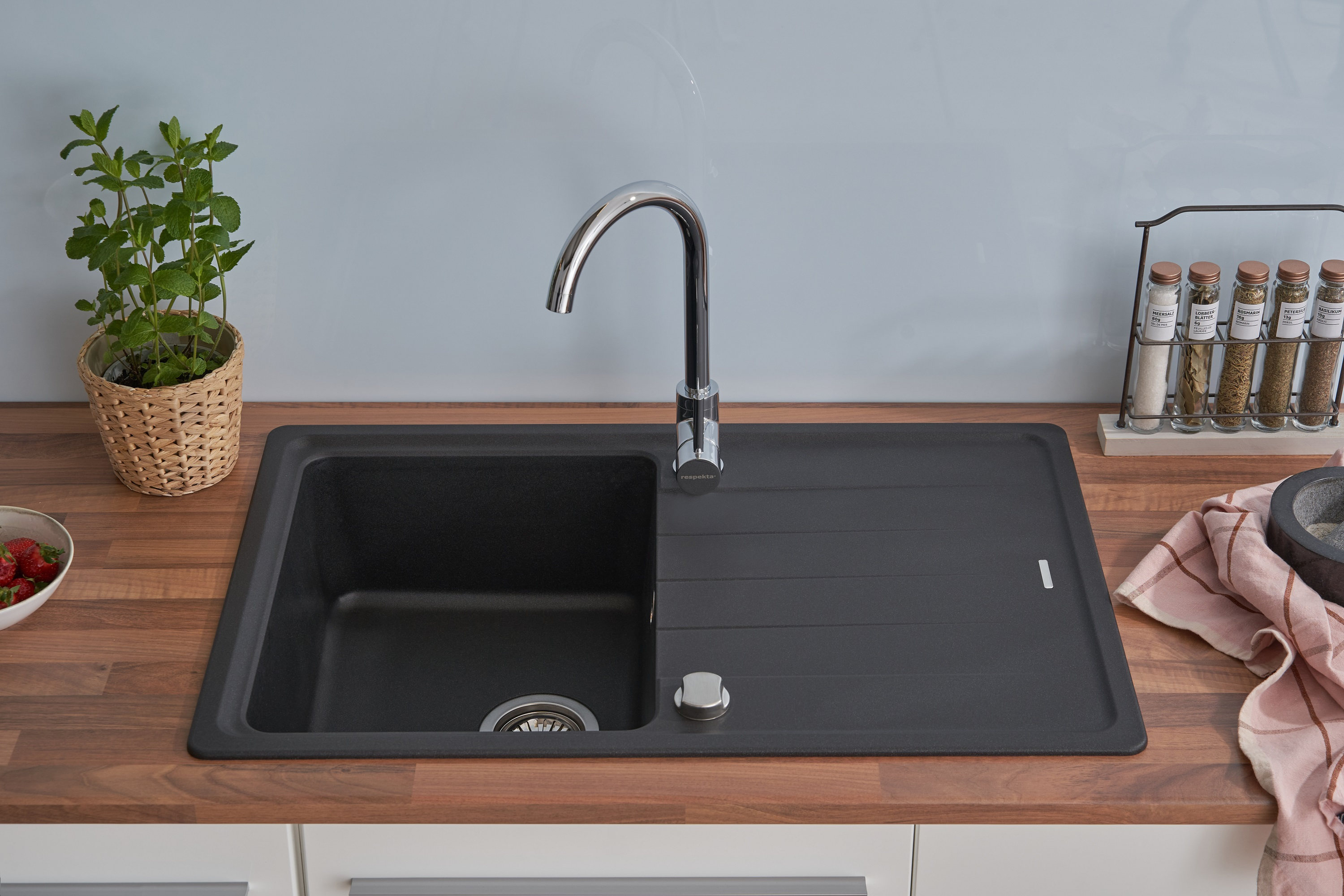 Schwarzes Waschbecken mit silbernem Wasserhahn. Grüne Pflanze und Erdbeeren. Hellblaue Wand im Hintergrund.
