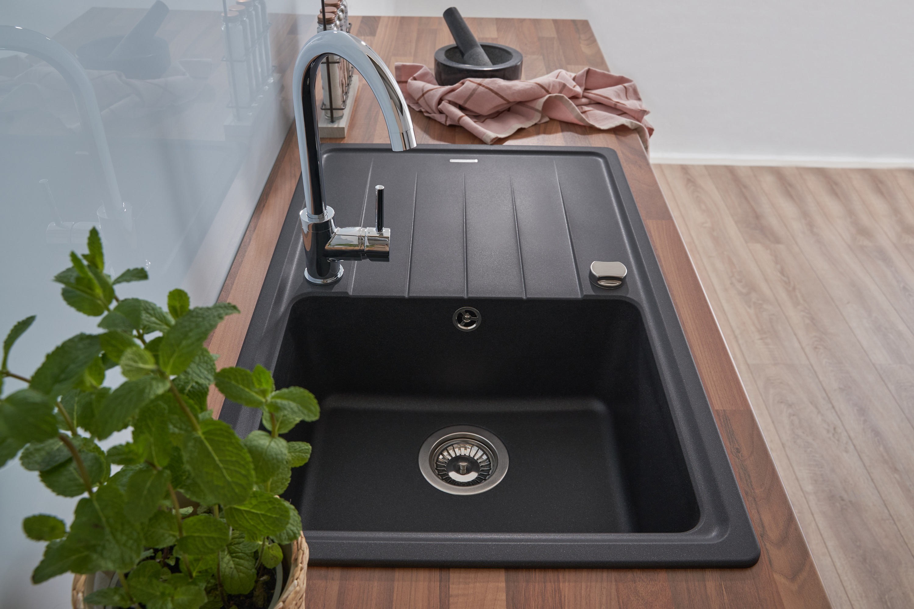 Schwarzes Waschbecken mit Chromarmatur. Holz-Arbeitsplatte mit grüner Pflanze und Mörser im Hintergrund.