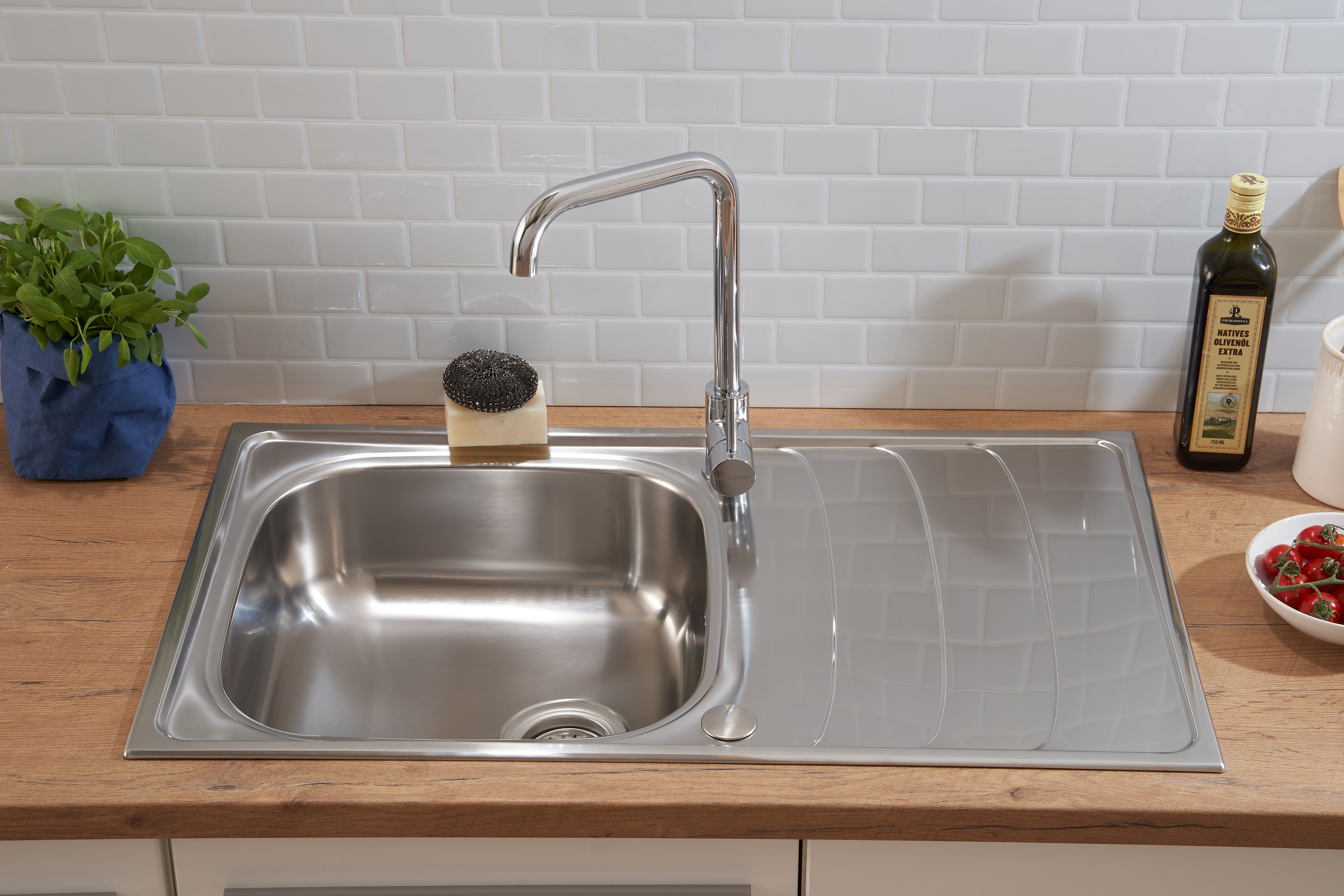 Ein Edelstahl-Küchenspülbecken mit Wasserhahn, Spülmittel und einer Schüssel mit Tomaten. Weiße Fliesen.