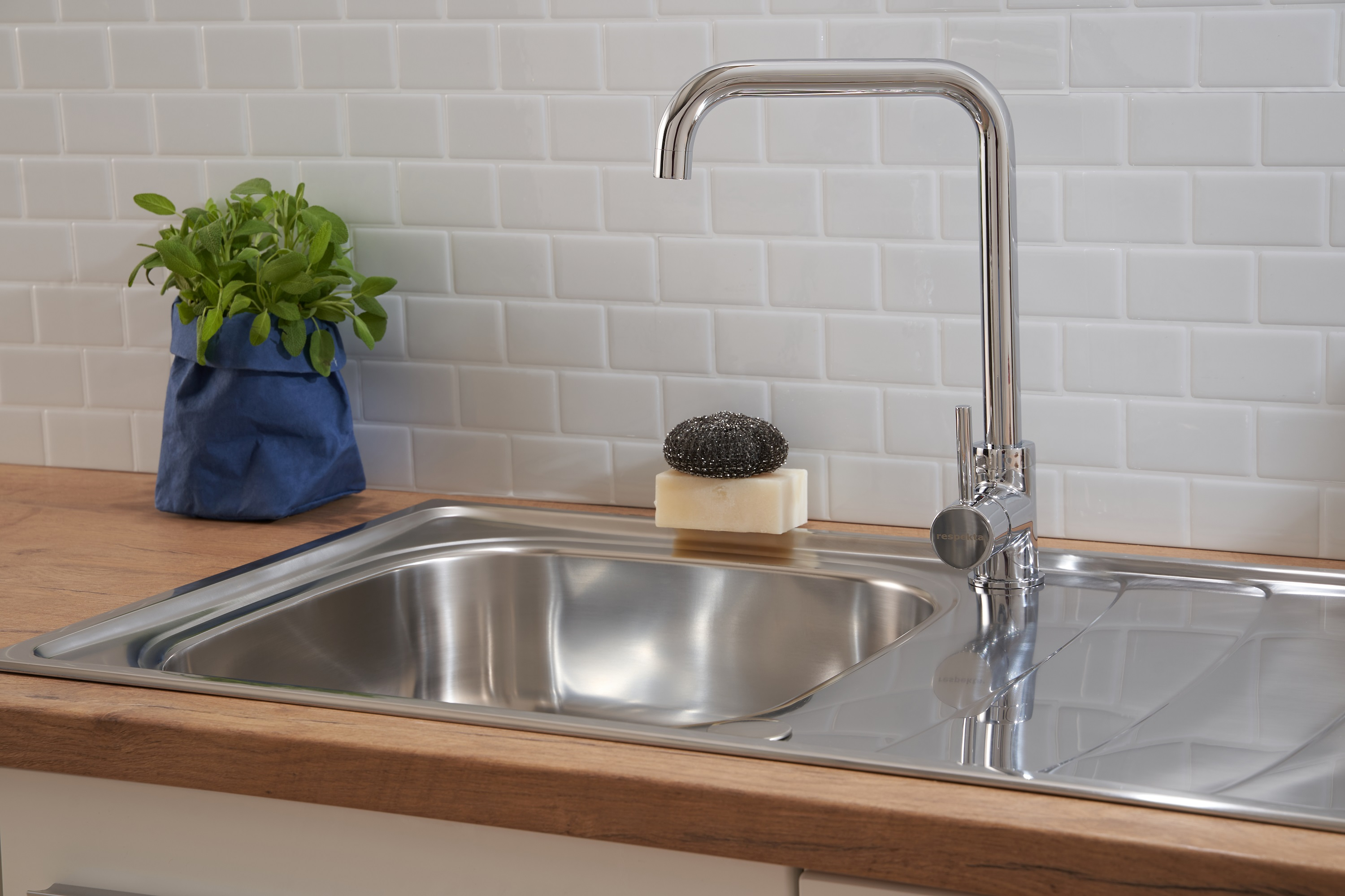 Ein Edelstahlbecken mit Wasserhahn. Eine blaue Papiertüte mit einer Pflanze.