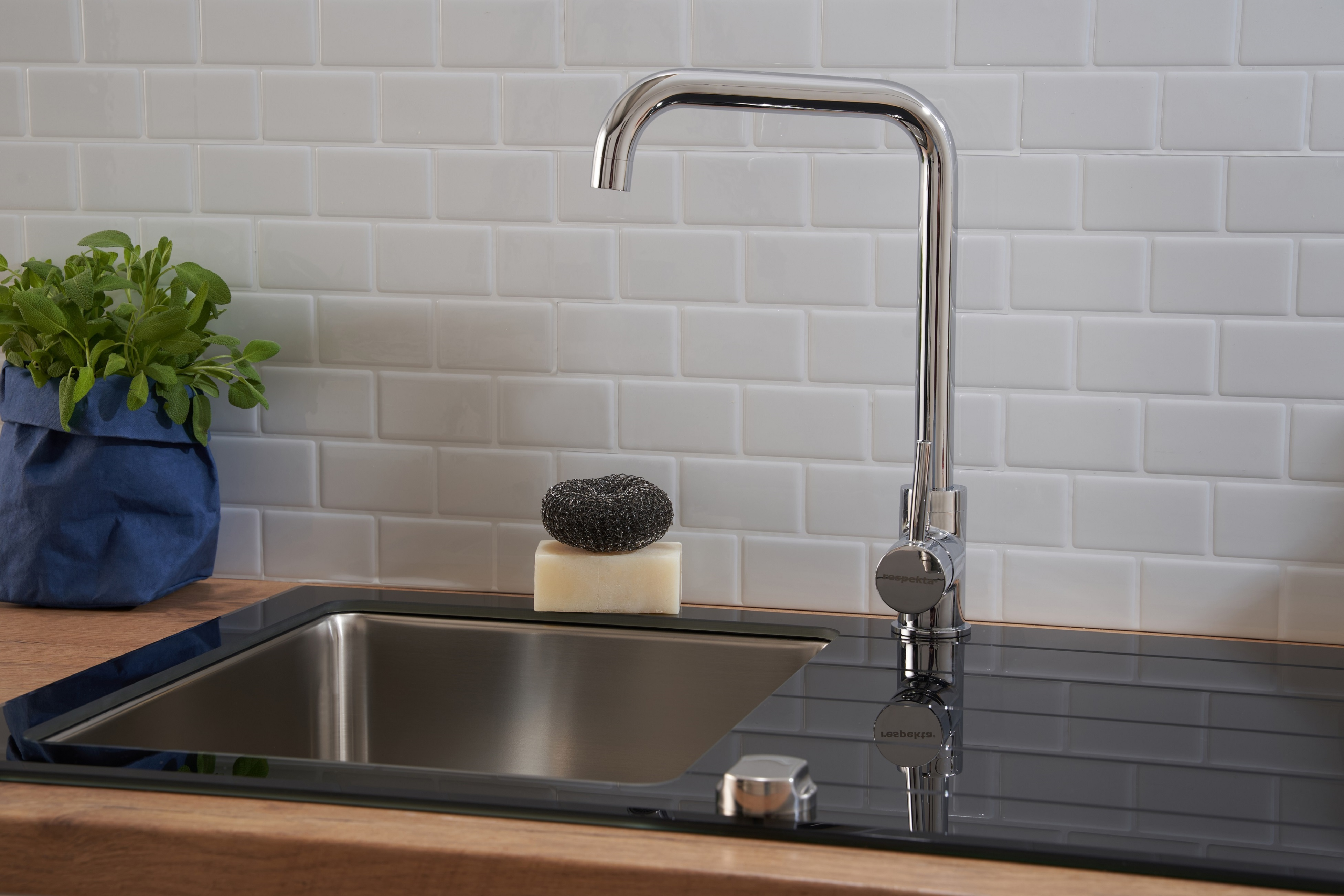 Küchenspüle mit Wasserhahn, Seife und Pflanze. Weiße Ziegelwand im Hintergrund.