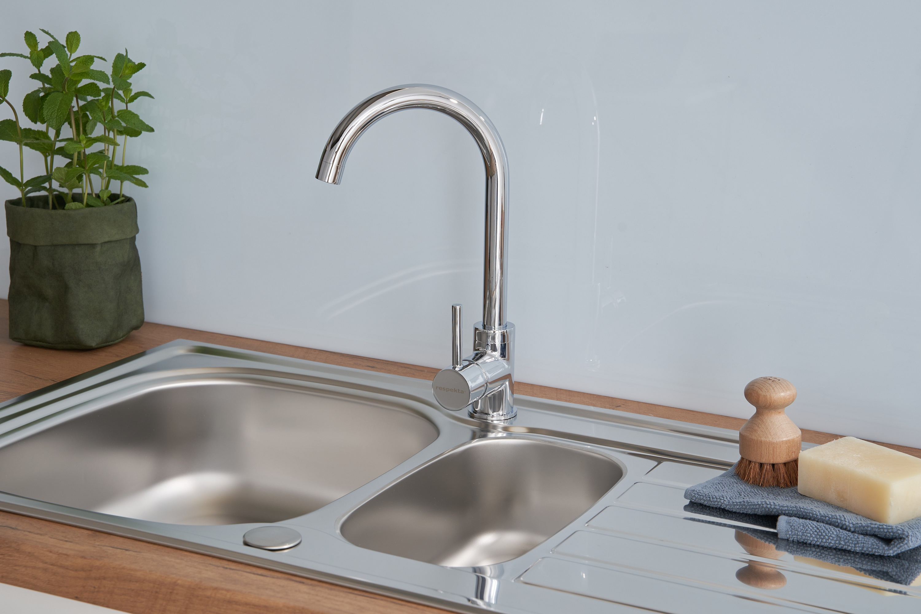 Edelstahlspüle mit Wasserhahn. Eine grüne Pflanze, Bürste und Seife sind auf der Theke sichtbar.