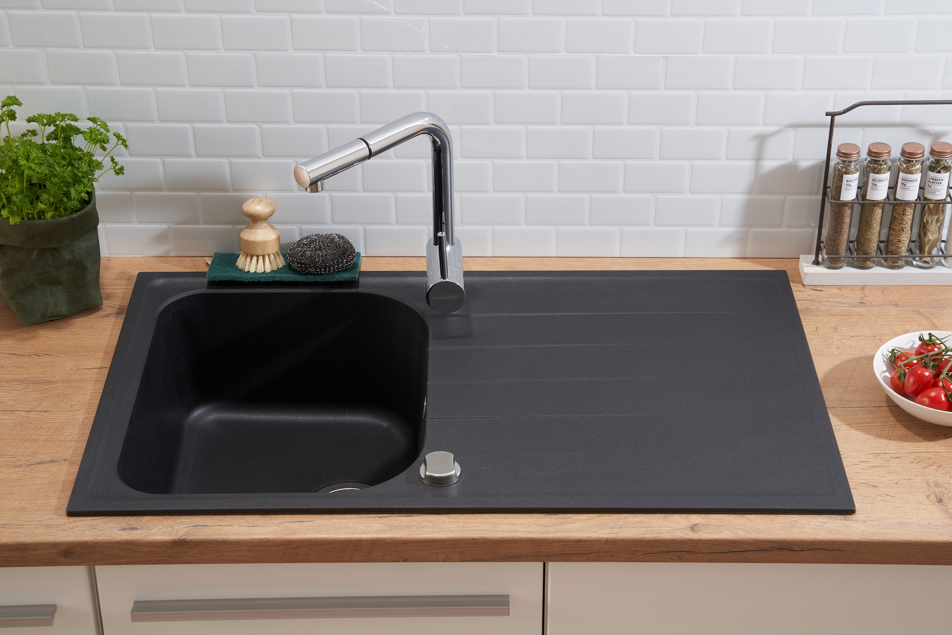 Ein Waschbecken mit Wasserhahn, Schwamm und Pflanze in einer blauen Tasche. Weiße Fliesenwand.