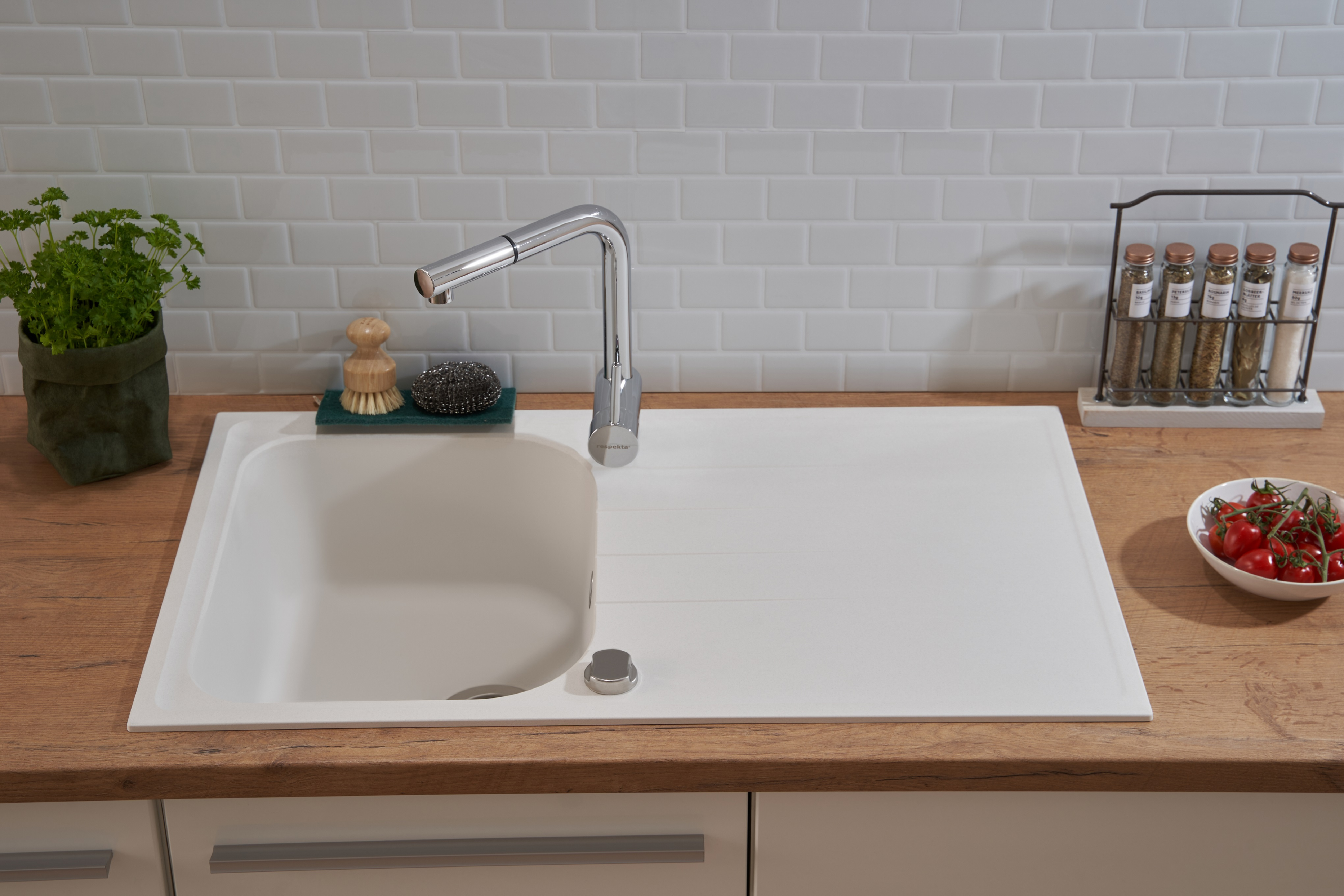Weißes Waschbecken mit Wasserhahn, eine kleine Schüssel mit Tomaten und eine Pflanze.
