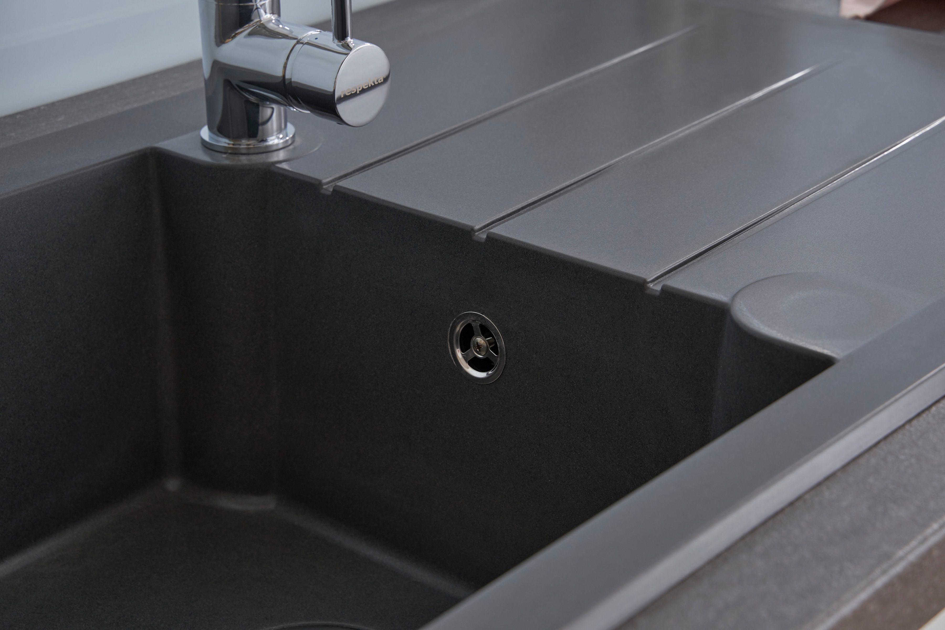 Graues Waschbecken mit Wasserhahn und Abfluss. Das Waschbecken hat ein eingebautes Abtropfbrett.