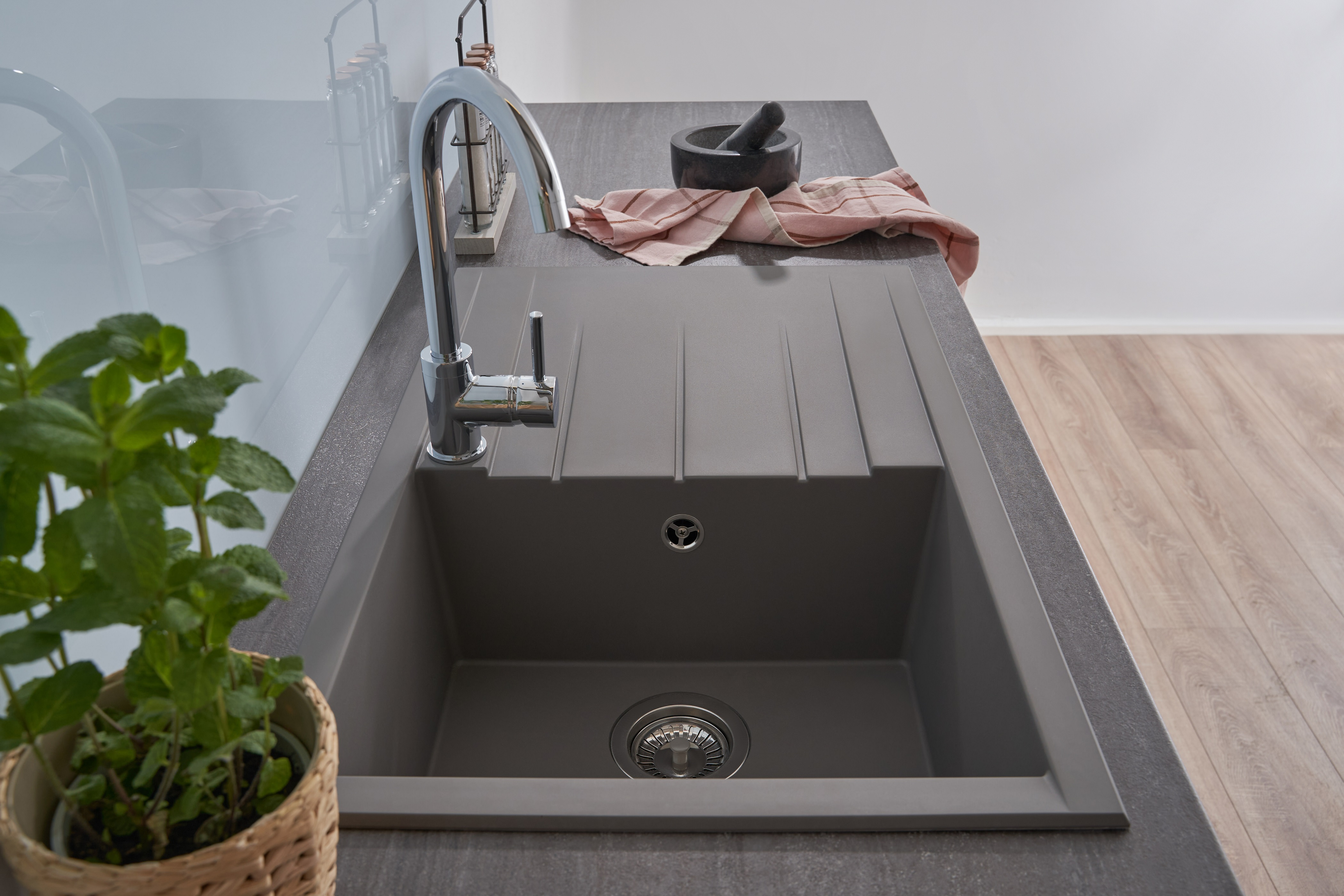 Graues Küchenbecken mit Wasserhahn. Minzpflanze im Korb links.