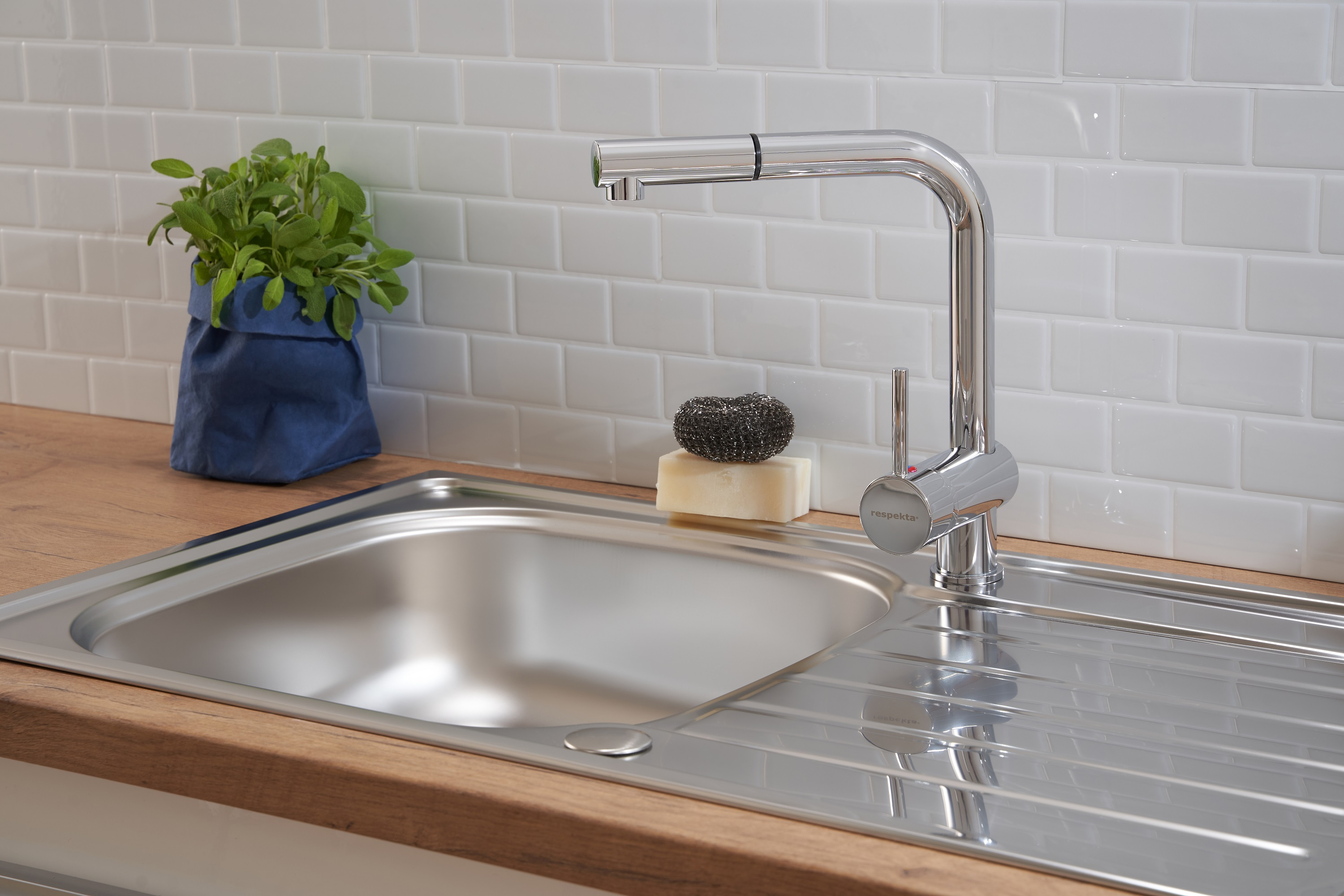 Küchenspüle mit Wasserhahn, Pflanze und Seife vor weißen Fliesen.