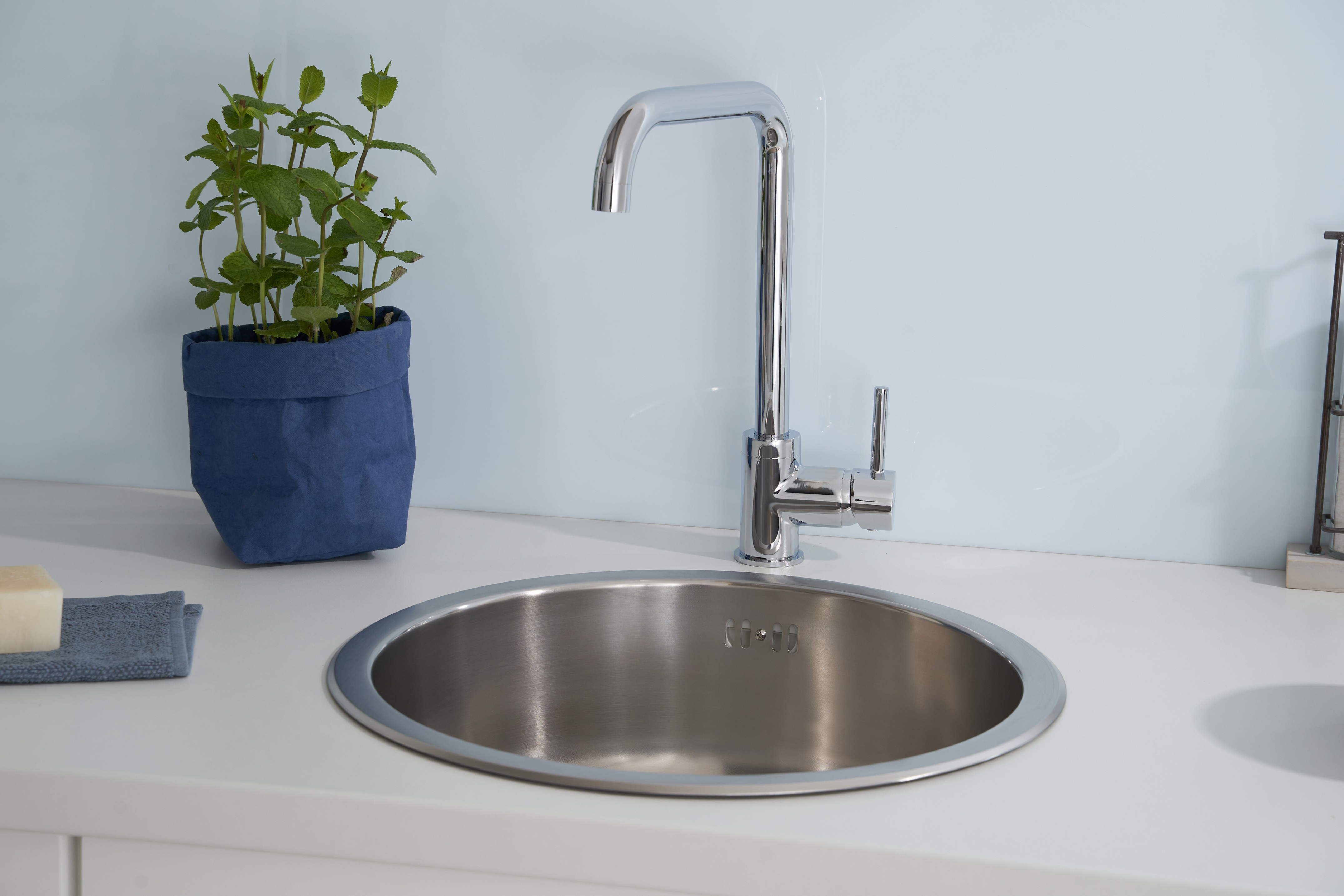 Edelstahlspüle mit hohem Wasserhahn, blauem Blumentopf und Seifenschale auf weißer Arbeitsplatte.