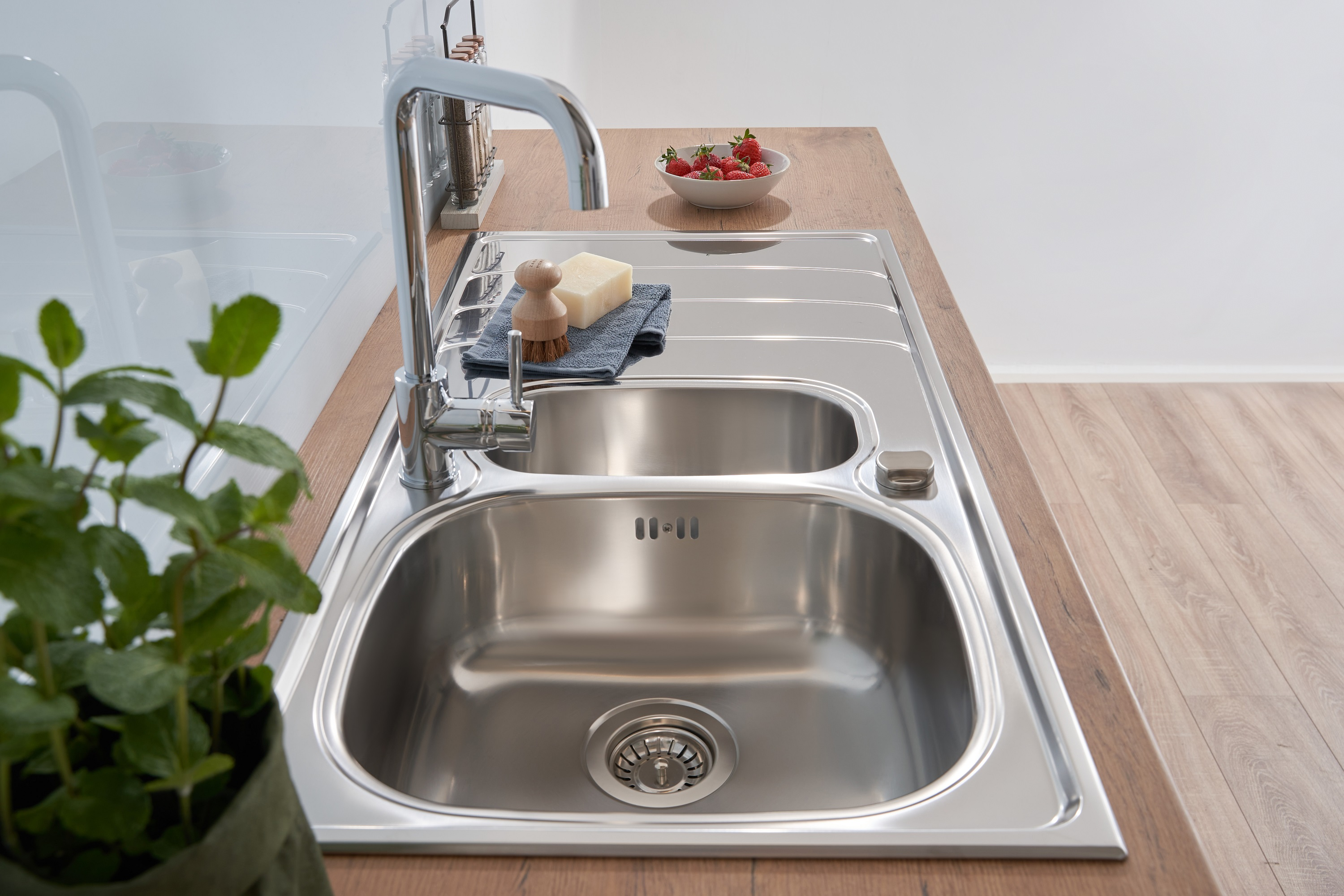 Küchenspüle mit Wasserhahn, Seife, Bürste und Erdbeeren. Holztheke und Pflanze in der Ecke.