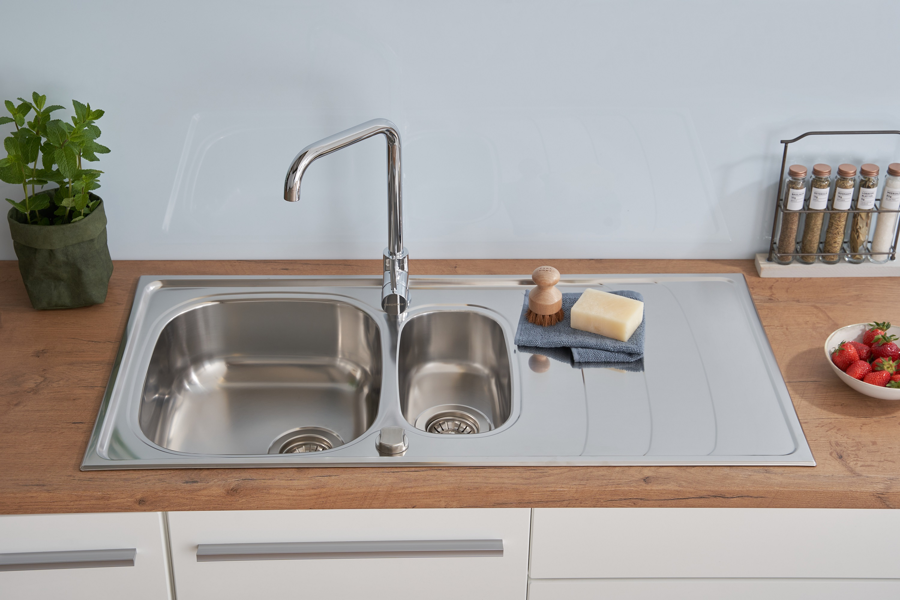 Edelstahl-Küchenspüle mit Wasserhahn, Seife, Schrubbbürste und Minzepflanze.