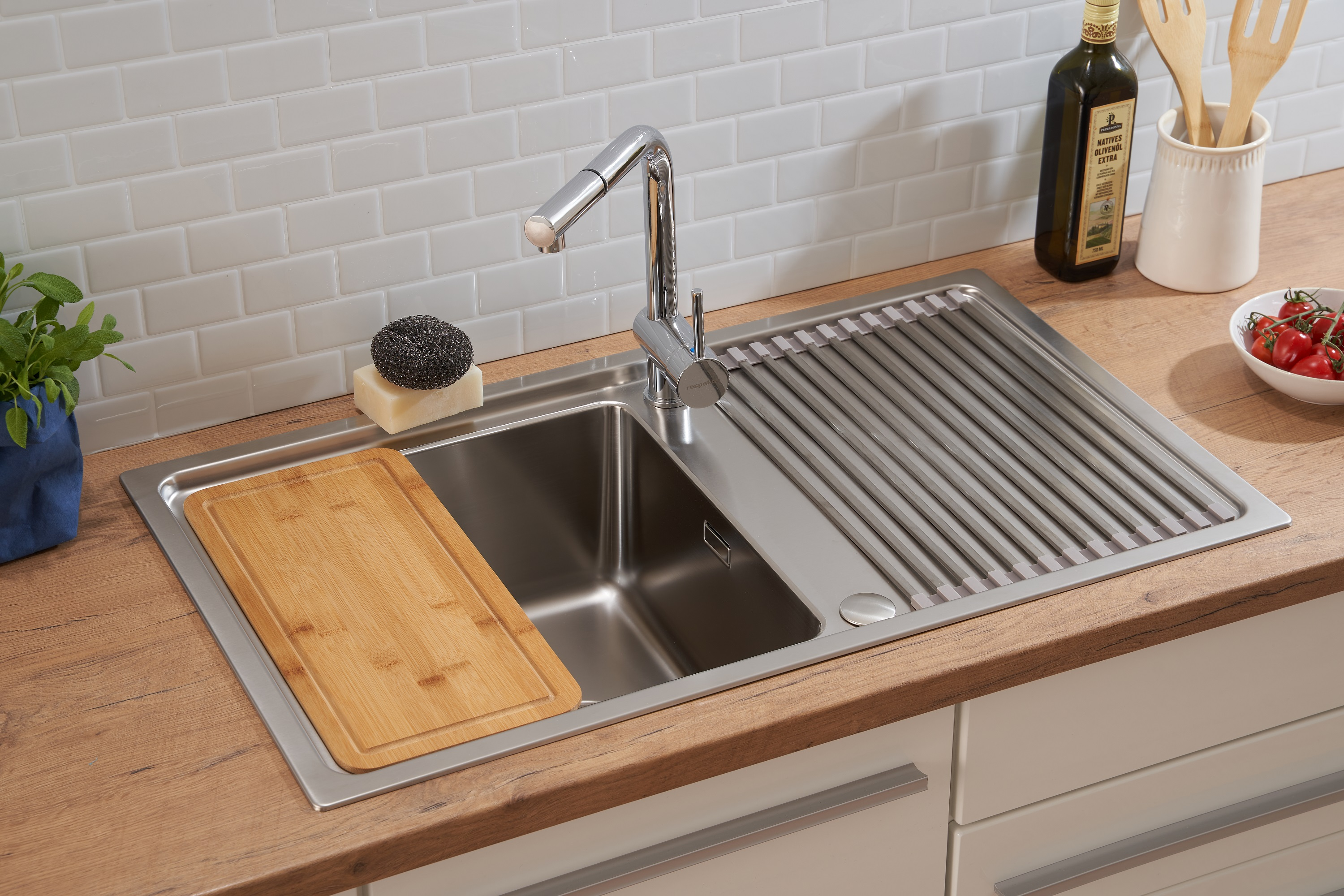Edelstahl-Küchenspüle mit Schneidebrett, Schwamm und Wasserhahn. Weiße Fliesenwand im Hintergrund, Olivenöl.