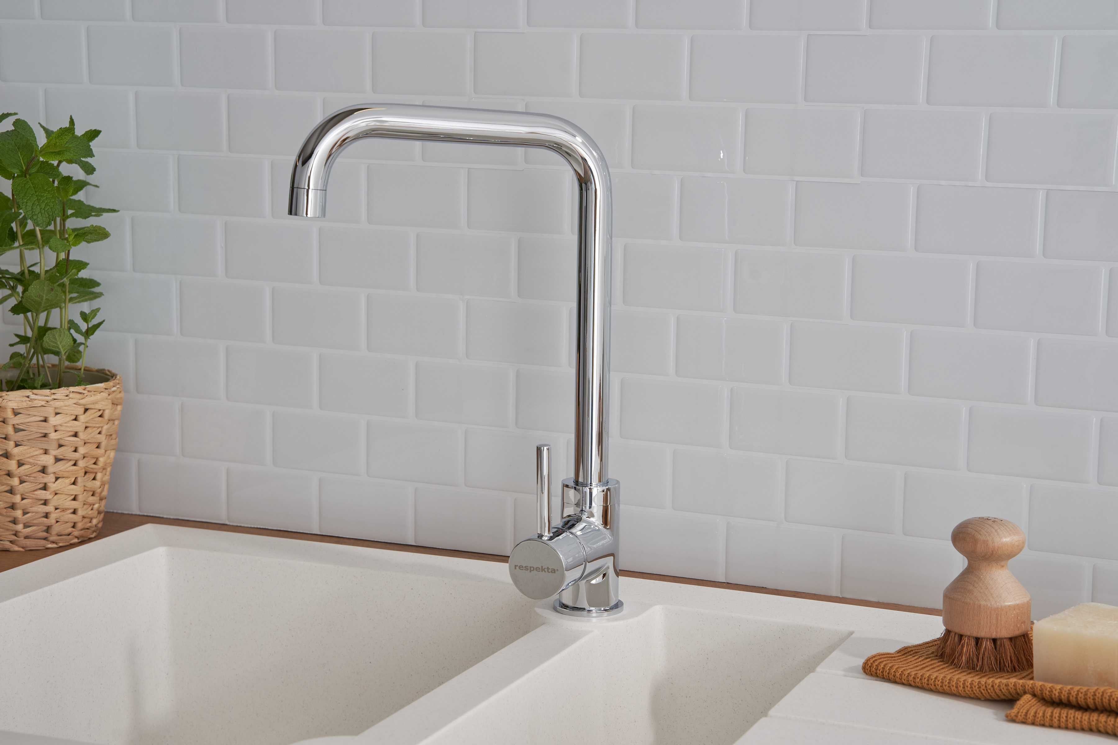 Ein verchromter Wasserhahn über einem weißen Waschbecken. Eine kleine Pflanze und eine Holzbürste stehen daneben.