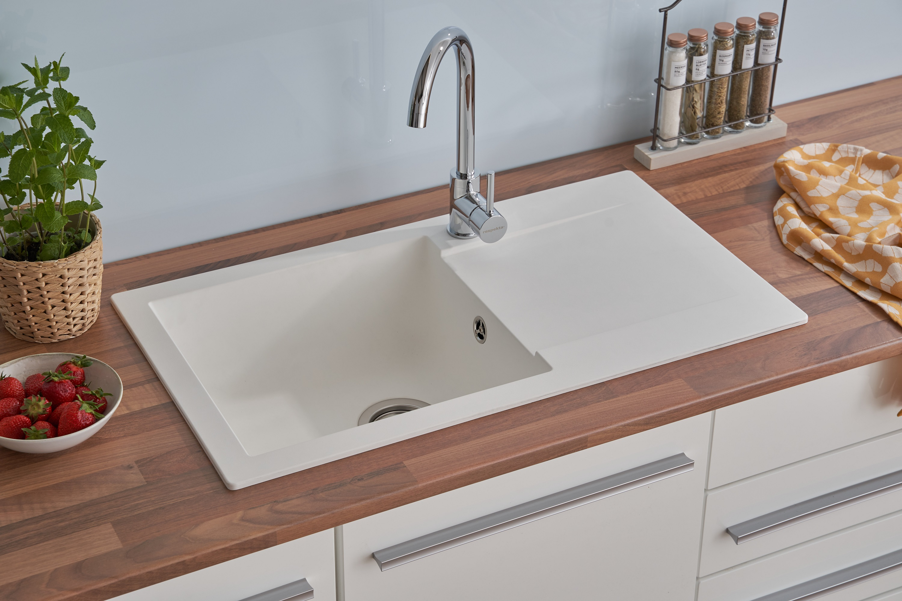 Ein weißes Waschbecken mit Chromarmatur, eine Holz-Arbeitsplatte und weiße Schränke. Erdbeeren sind in einer Schüssel.