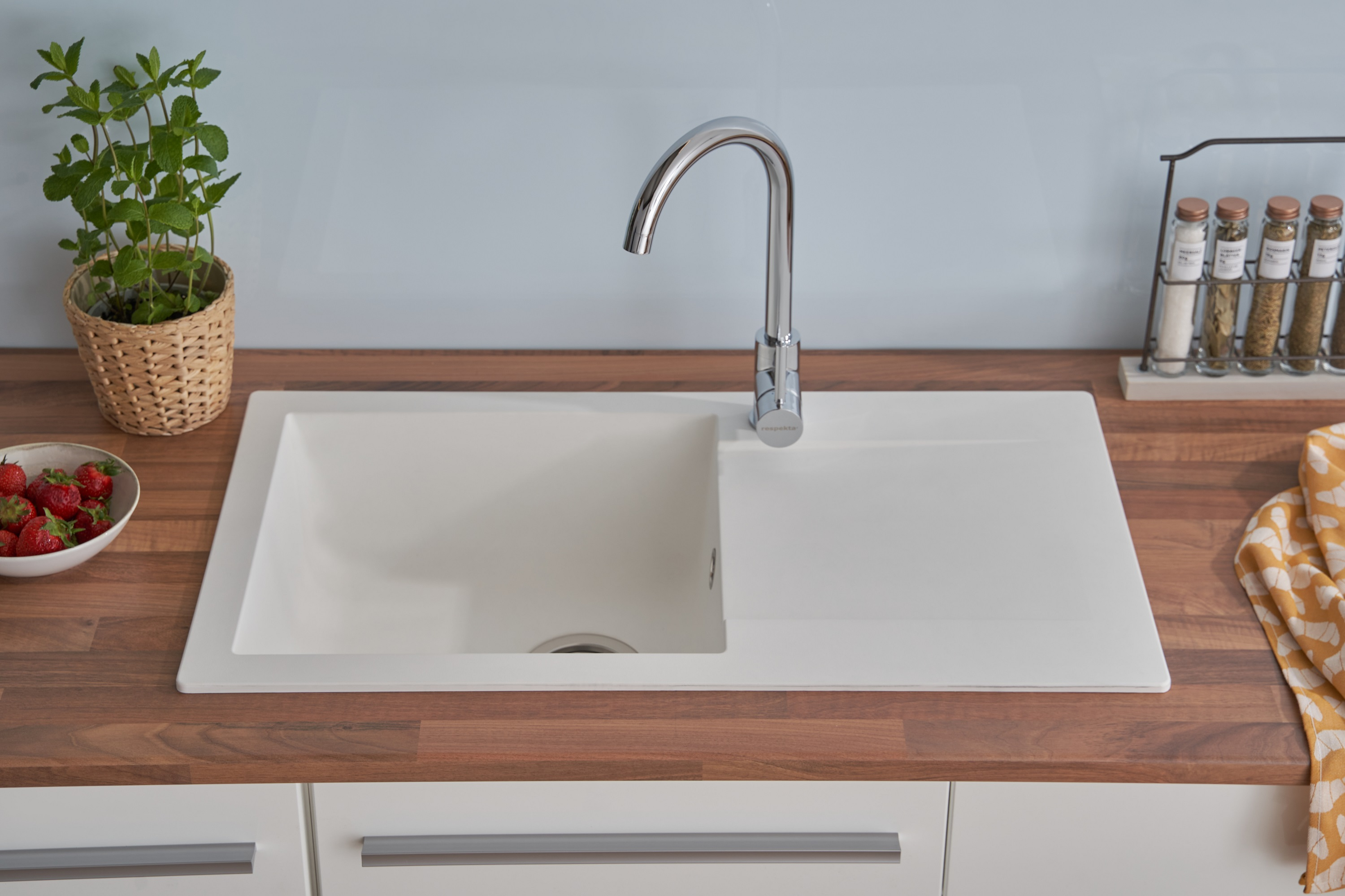 Weißes Küchenbecken mit Wasserhahn. Erdbeeren in Schale und Pflanze sichtbar.