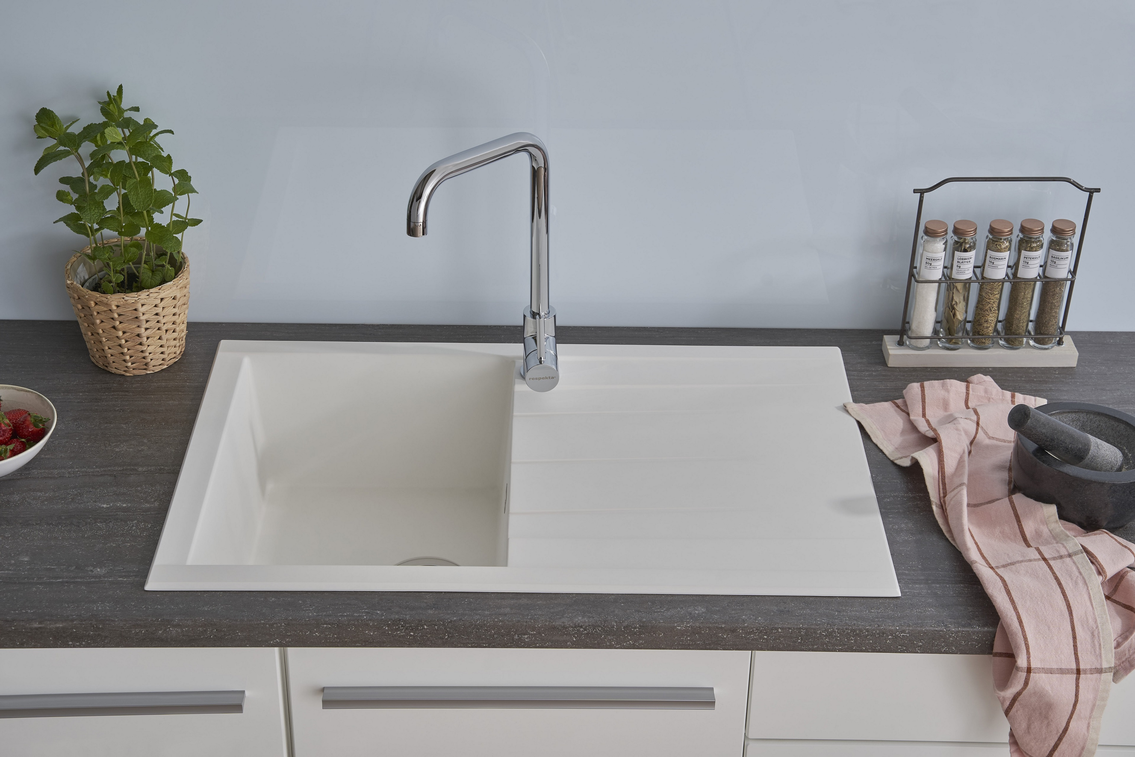 Ein weißes Küchenbecken mit Wasserhahn, Kräutertopf, Gewürzen und einem Geschirrtuch.