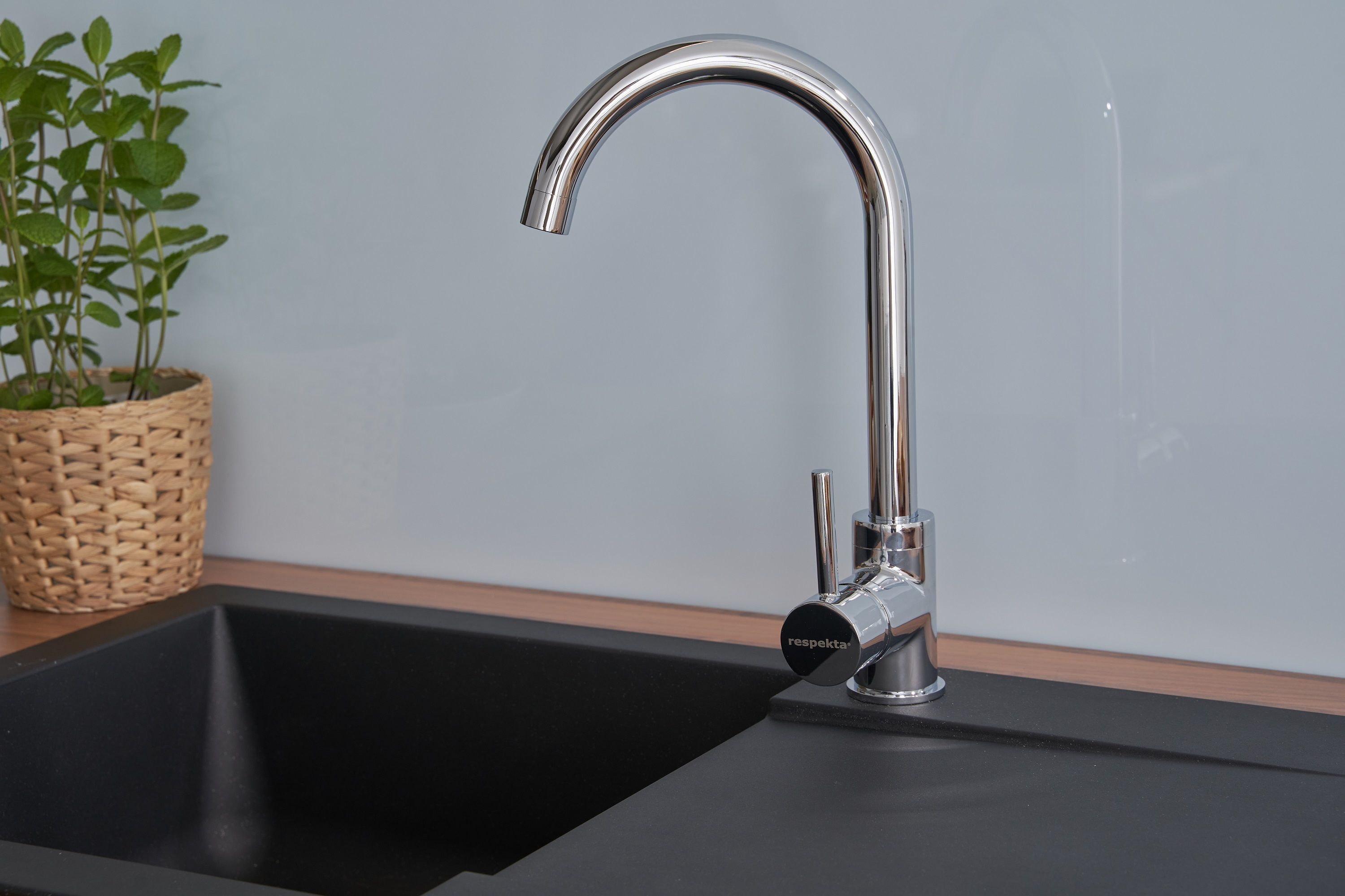 Chrom-Küchenarmatur über schwarzem Waschbecken, mit einem Weidenkorb mit Minze in der Ecke.