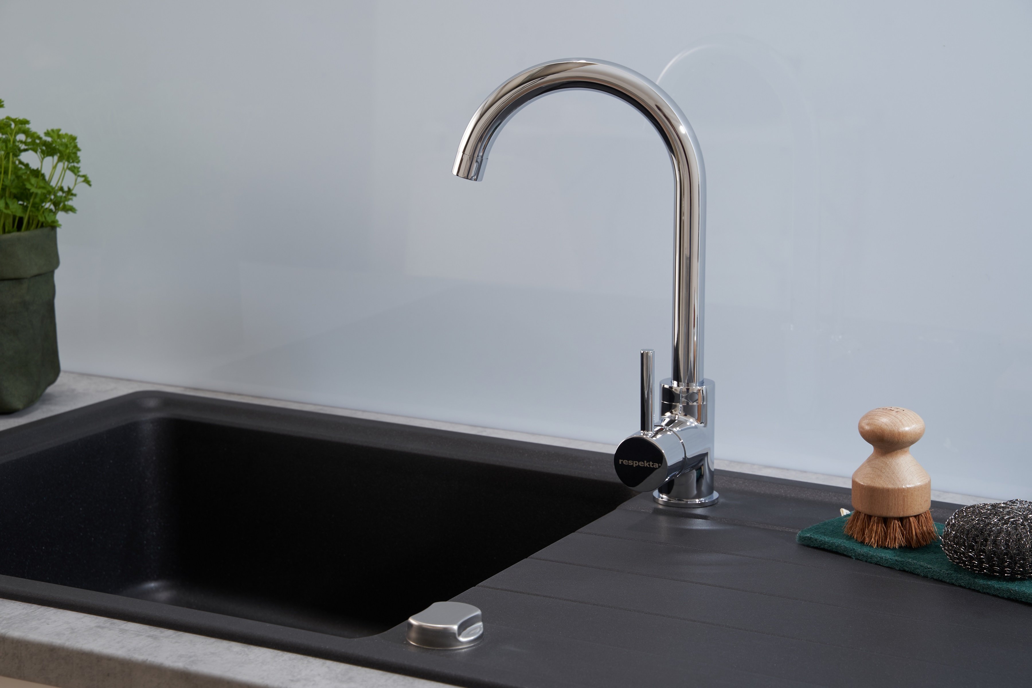 Ein verchromter Wasserhahn über einem schwarzen Waschbecken in einer Küche. Eine grüne Pflanze und ein Schwamm auf der Theke.