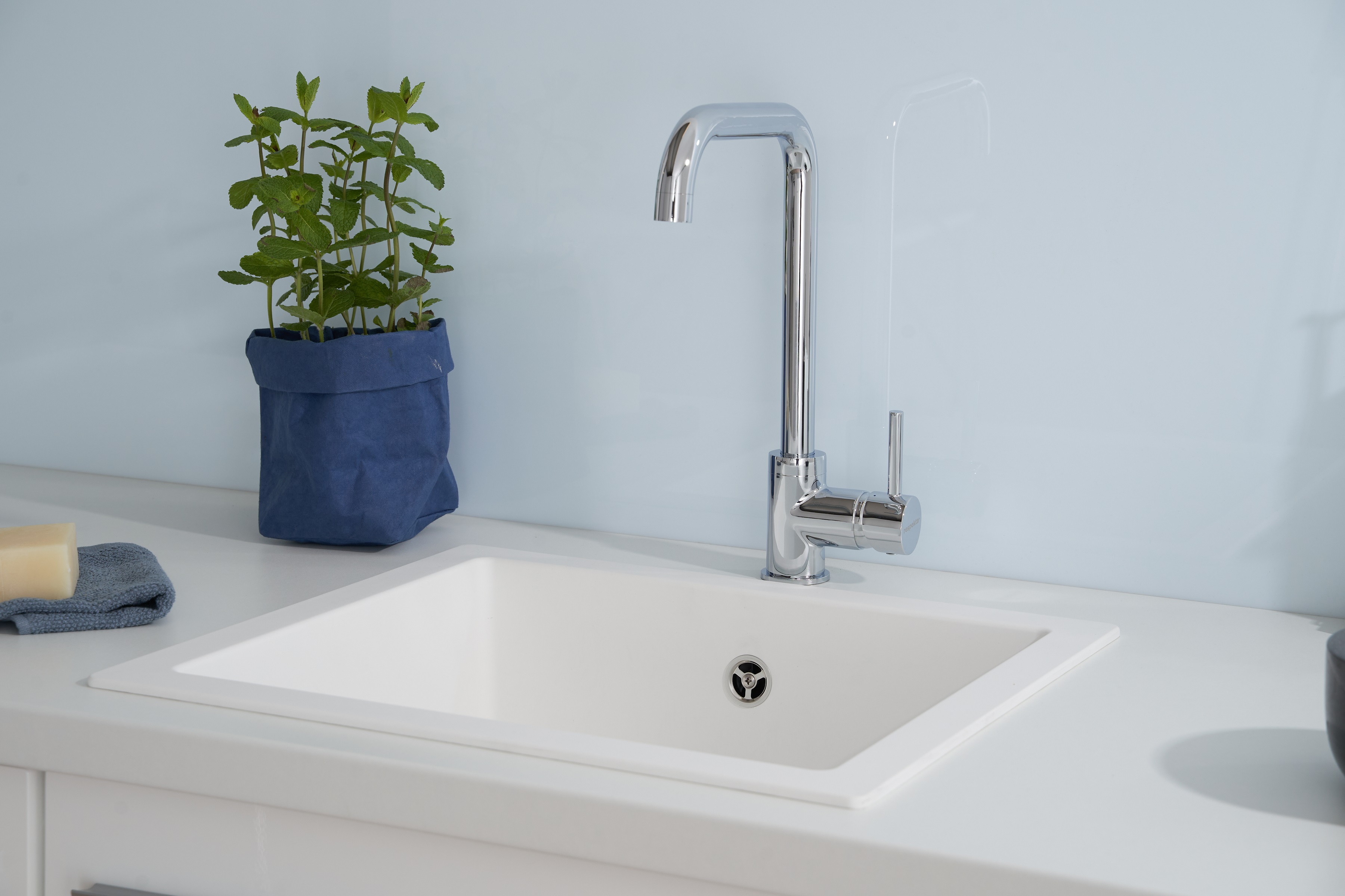 Weißes Küchenbecken mit Wasserhahn und einer Pflanze in einem blauen Topf links.