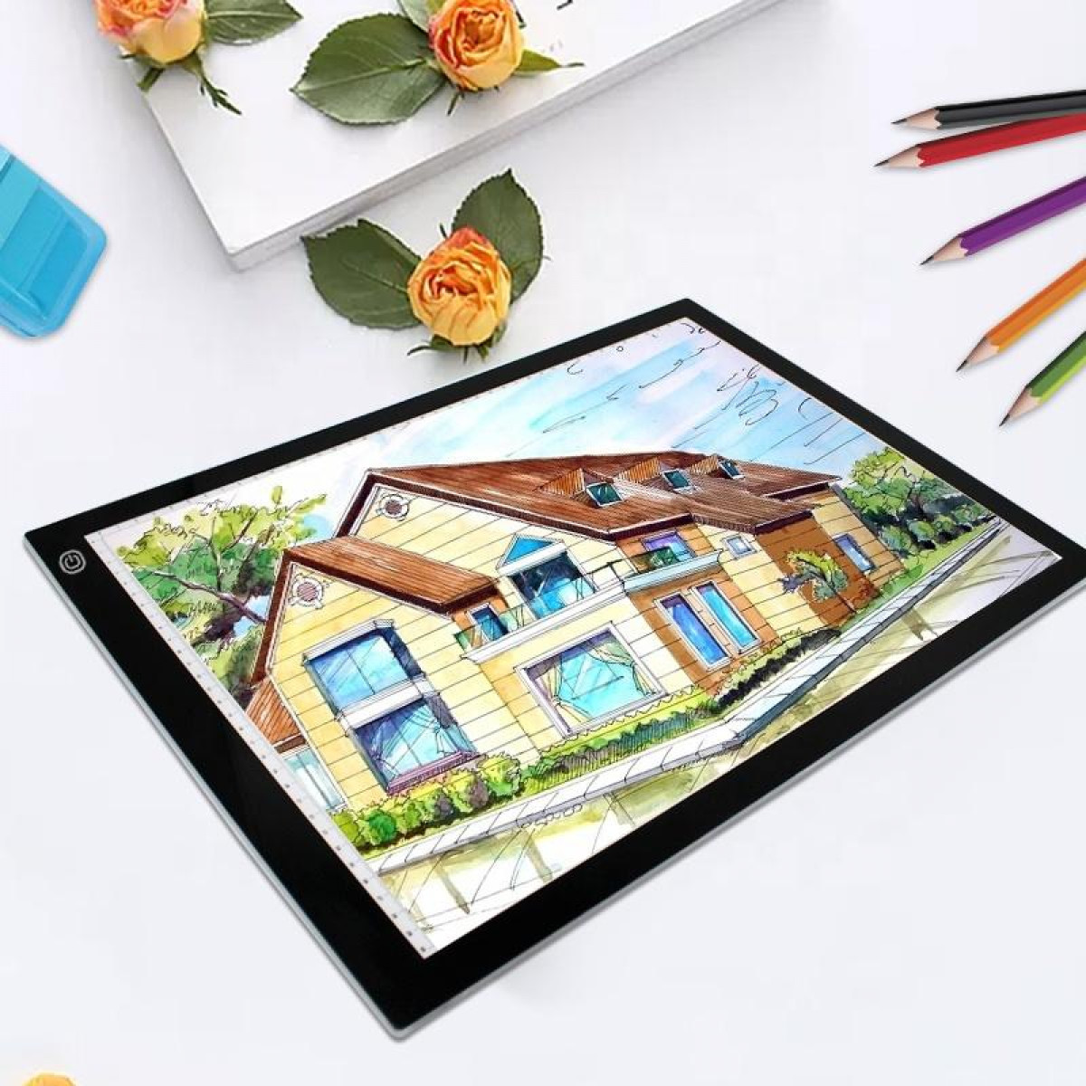 Stół świetlny z rysunkiem domu, ołówkami i kwiatami. Stół stoi na białej powierzchni.