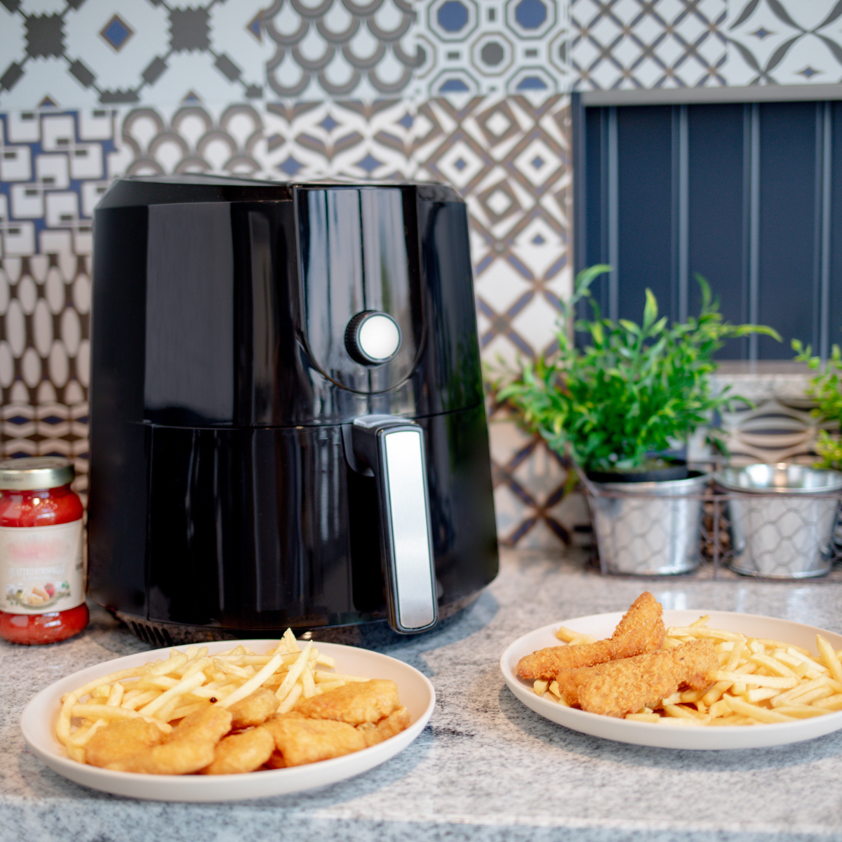 Schwarze Heißluftfritteuse mit Essen. Pommes und Chicken Nuggets auf Tellern. Ein Glas Soße.