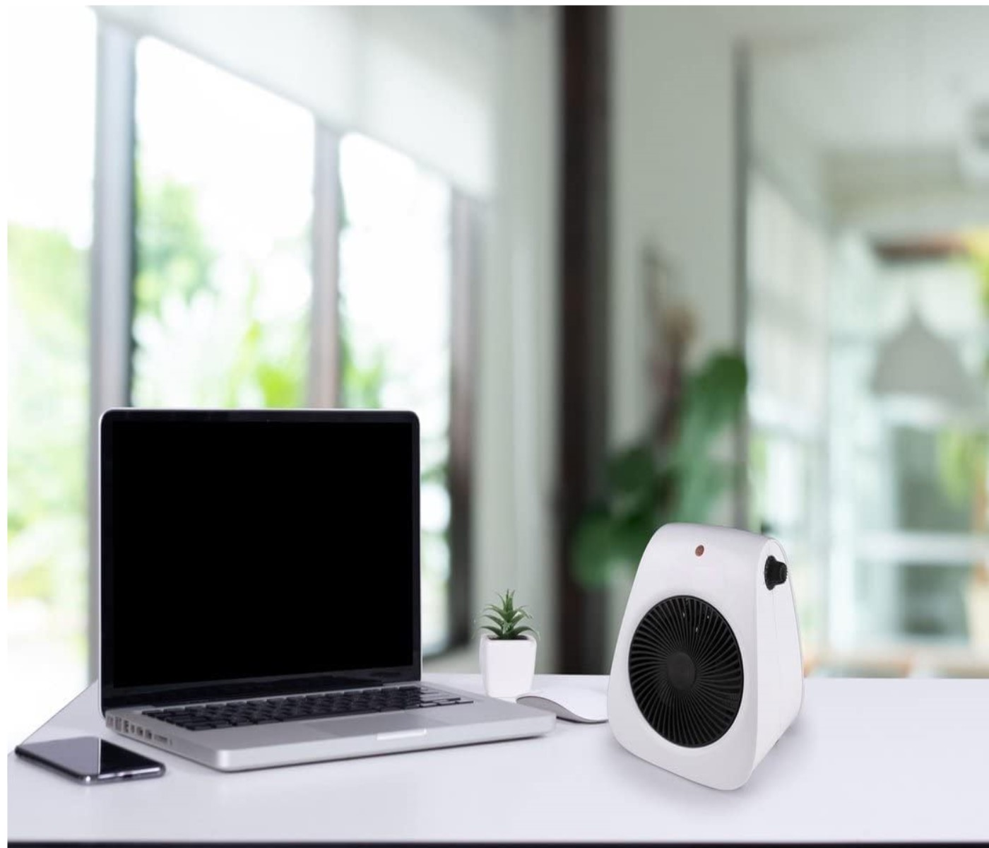 Ein Laptop, eine kleine weiße Heizung und ein Telefon stehen auf einem weißen Schreibtisch. Ein Fenster ist im Hintergrund.