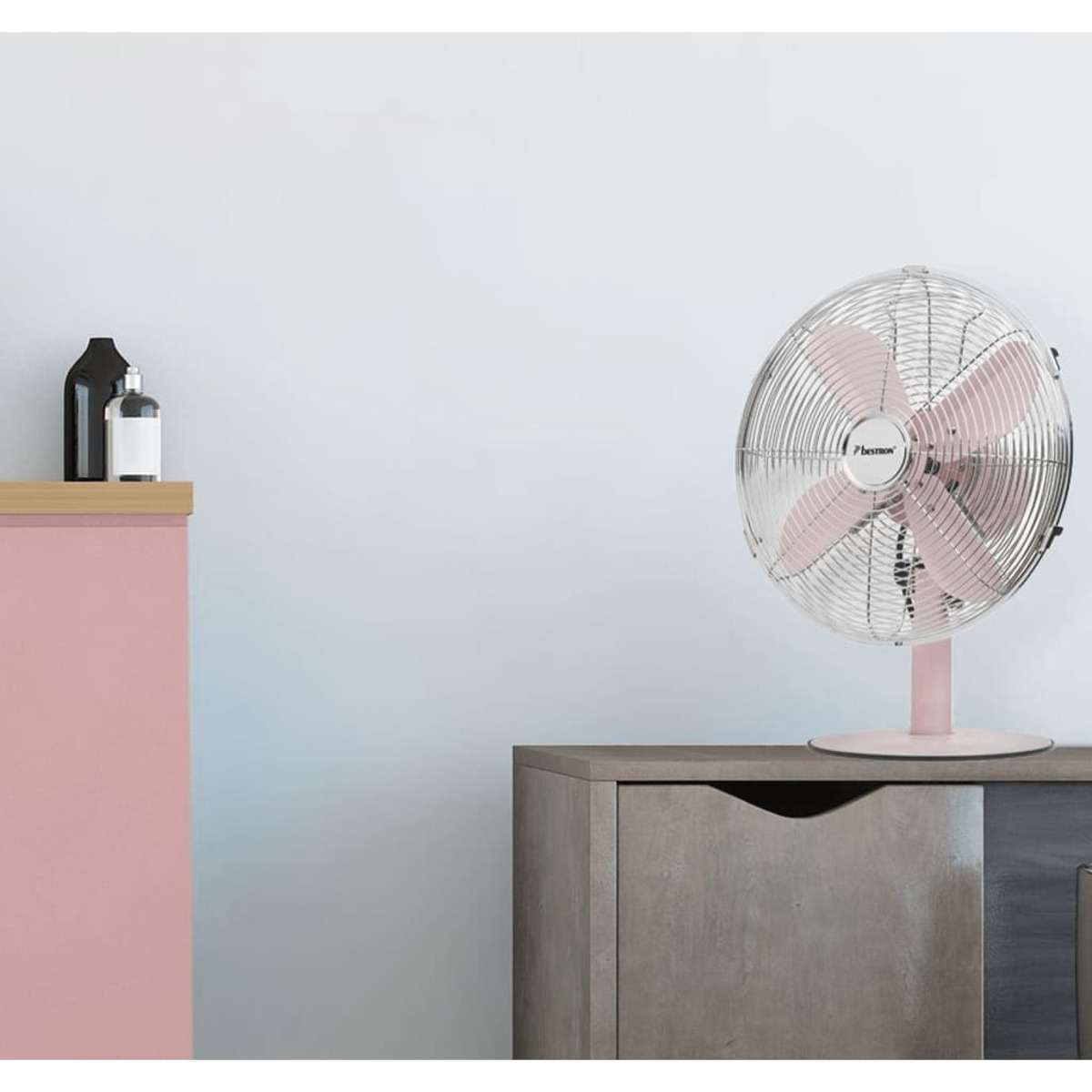 Un ventilatore rosa su un supporto rosa, posizionato su un mobile grigio contro un muro bianco.