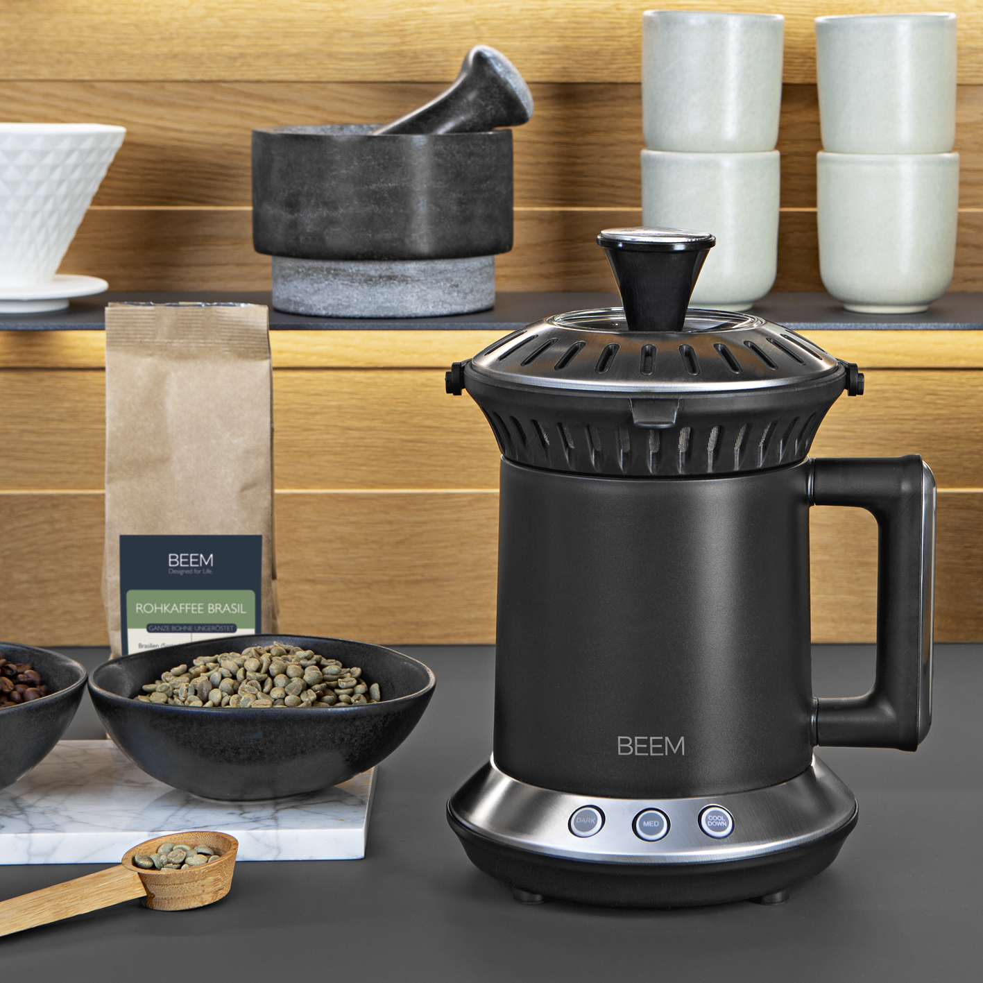 Schwarze Kaffeemaschine mit Griff und silberner Basis. Bohnen, Tassen und Holzlöffel sichtbar.