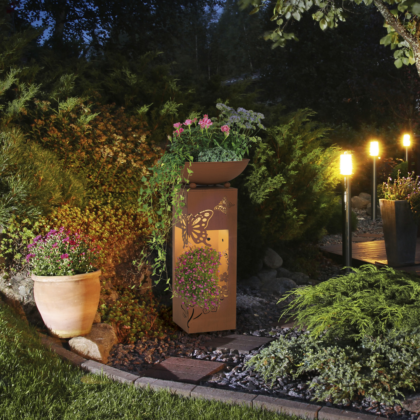 Gartenlandschaft im Freien bei Nacht. Metallskulptur mit Blumen und Lichtern, von Büschen umgeben.