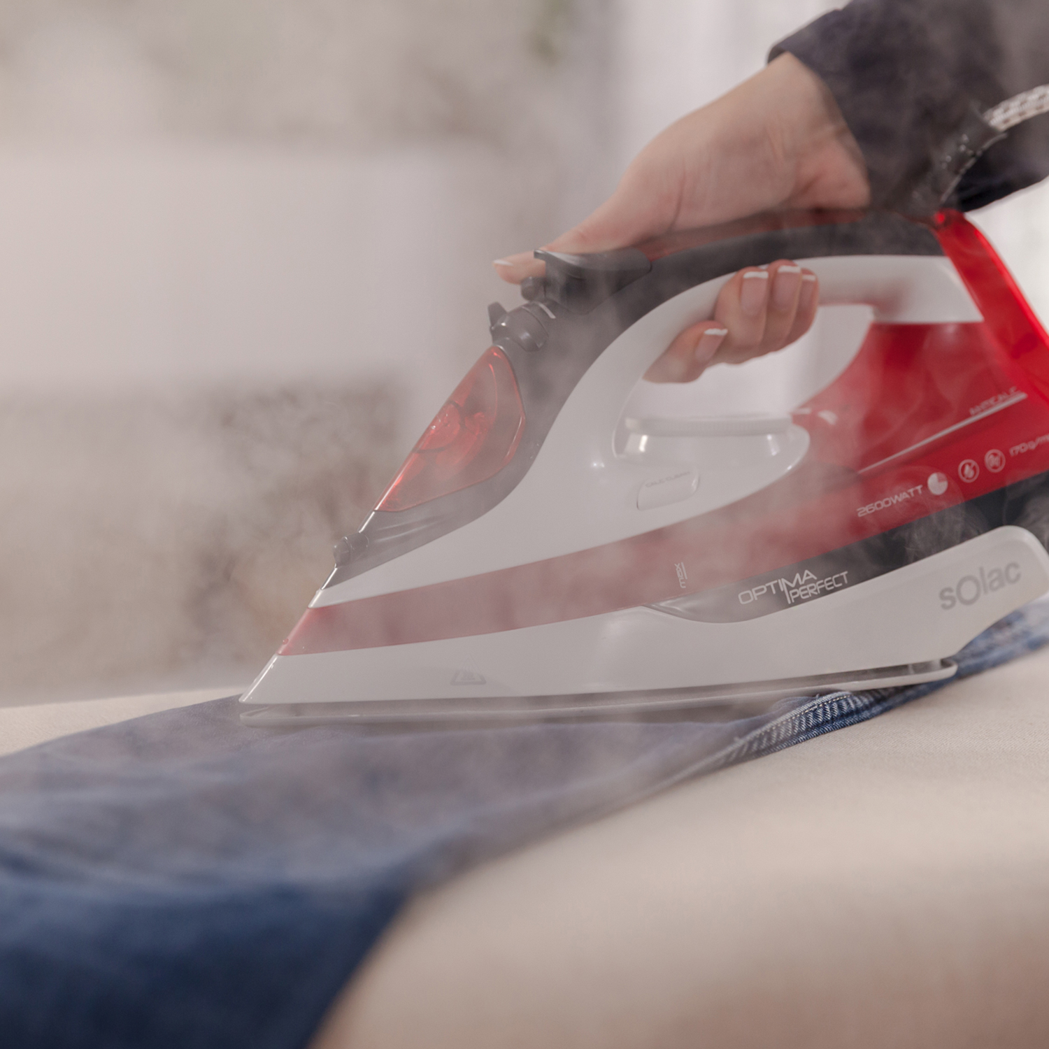 Una persona planchando vaqueros azules. Plancha roja y blanca, vapor y una superficie blanca.