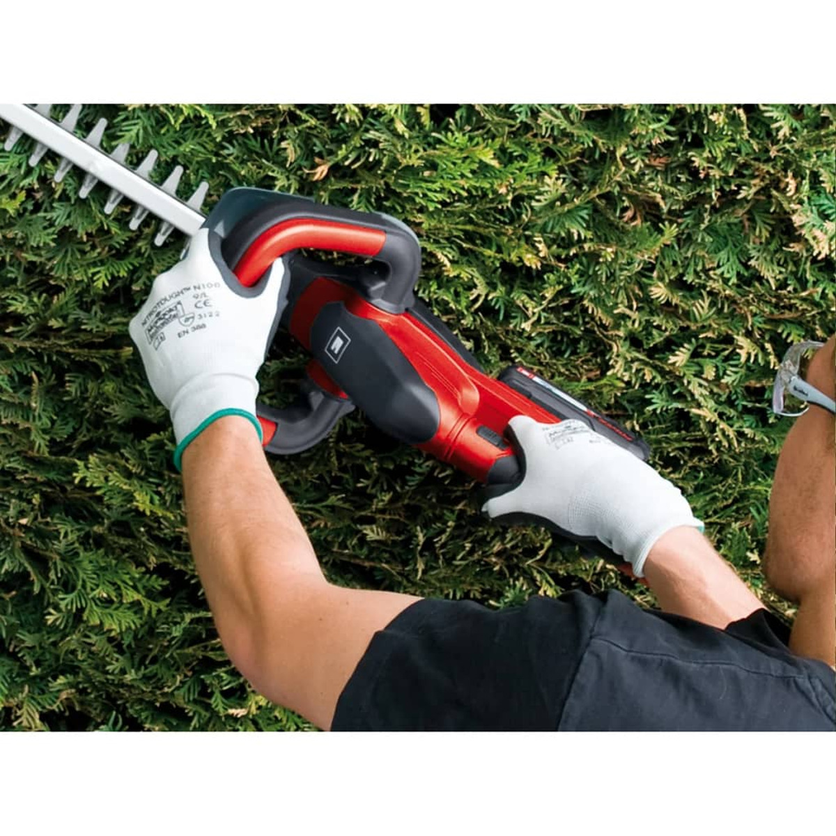 Osoba używająca nożyc do żywopłotu na zielonym żywopłocie. Noszą białe rękawice.