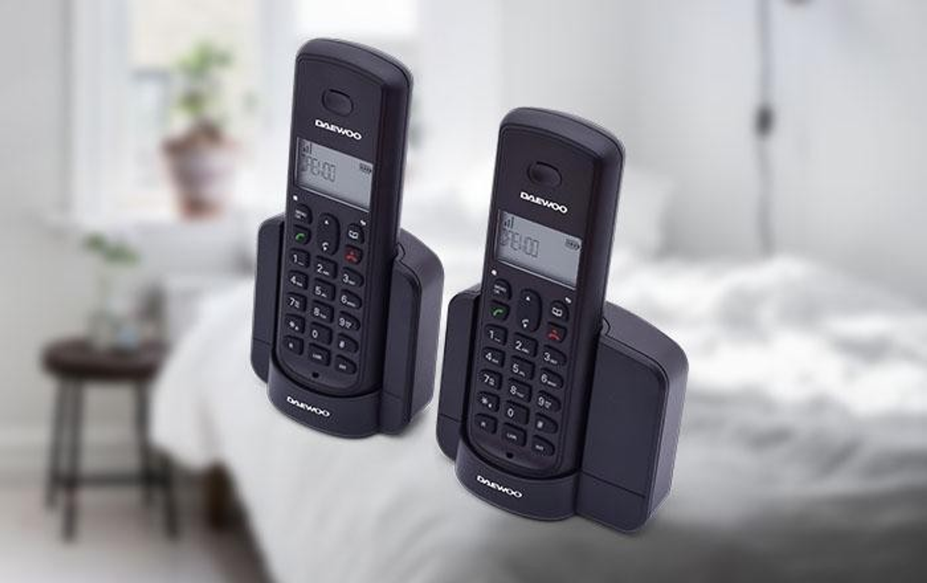 Dos teléfonos inalámbricos negros en sus estaciones de carga. Una cama y una pared blanca están en el fondo. Los teléfonos son Daewoo.