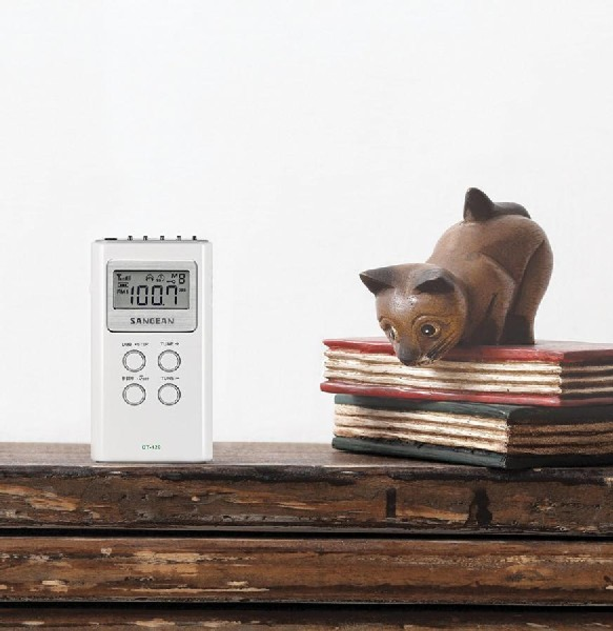 Ein weißes tragbares Radio mit Display, das Zahlen zeigt, neben einer Holzkatzenfigur auf Büchern.