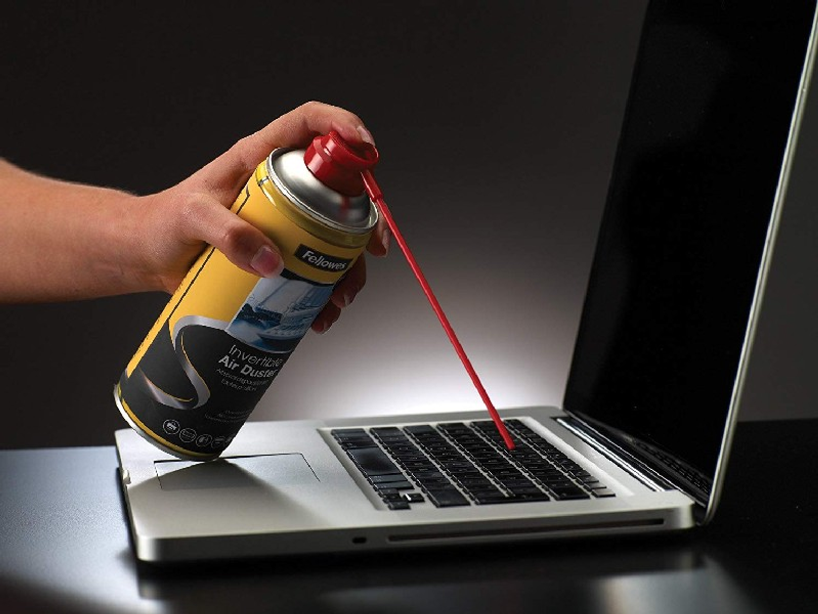 Ręka spryskuje klawiaturę laptopa sprężonym powietrzem. Puszka jest żółta i oznaczona 'Air Duster'.