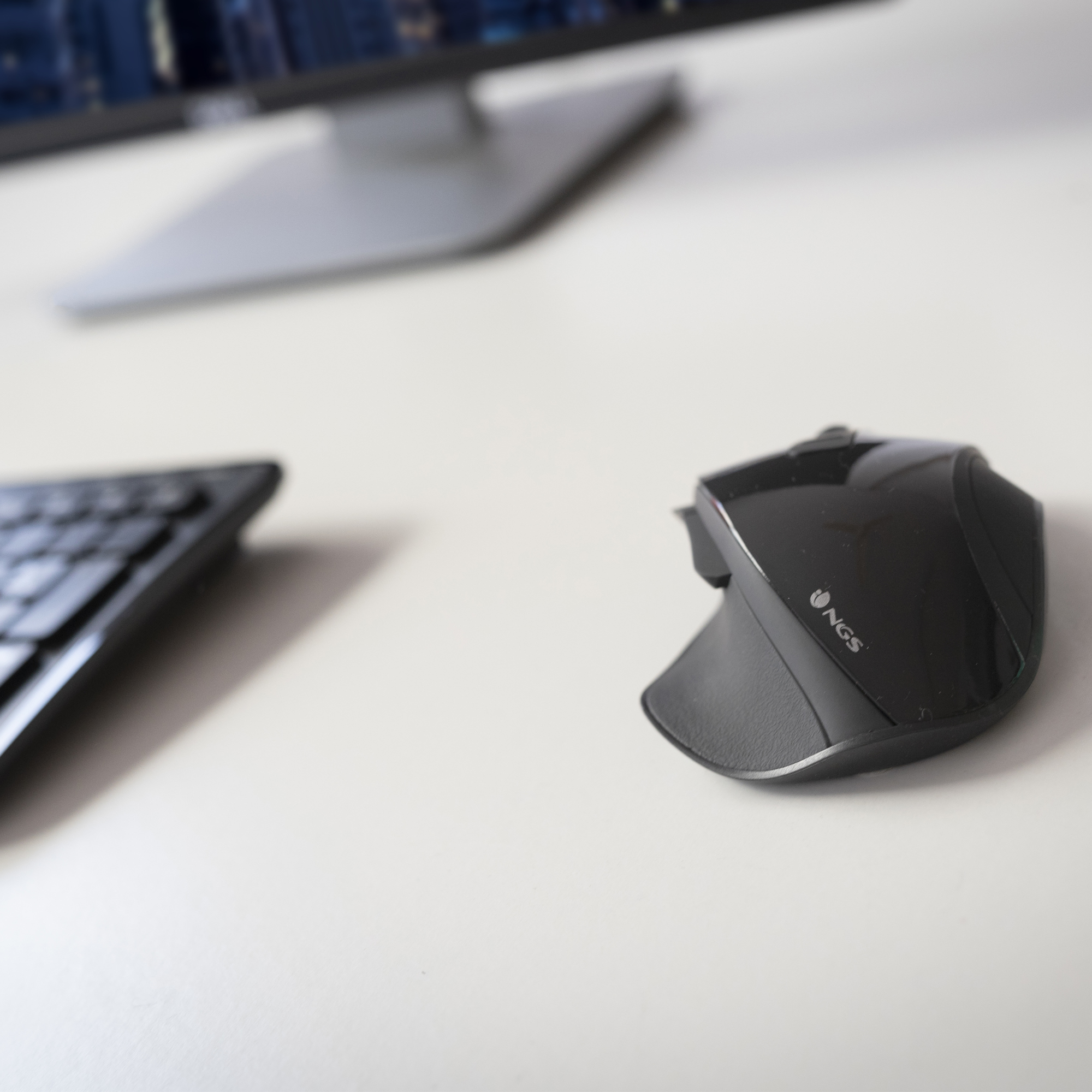Schwarze NGS-Maus auf weißer Oberfläche, Tastatur und Monitor im Hintergrund.