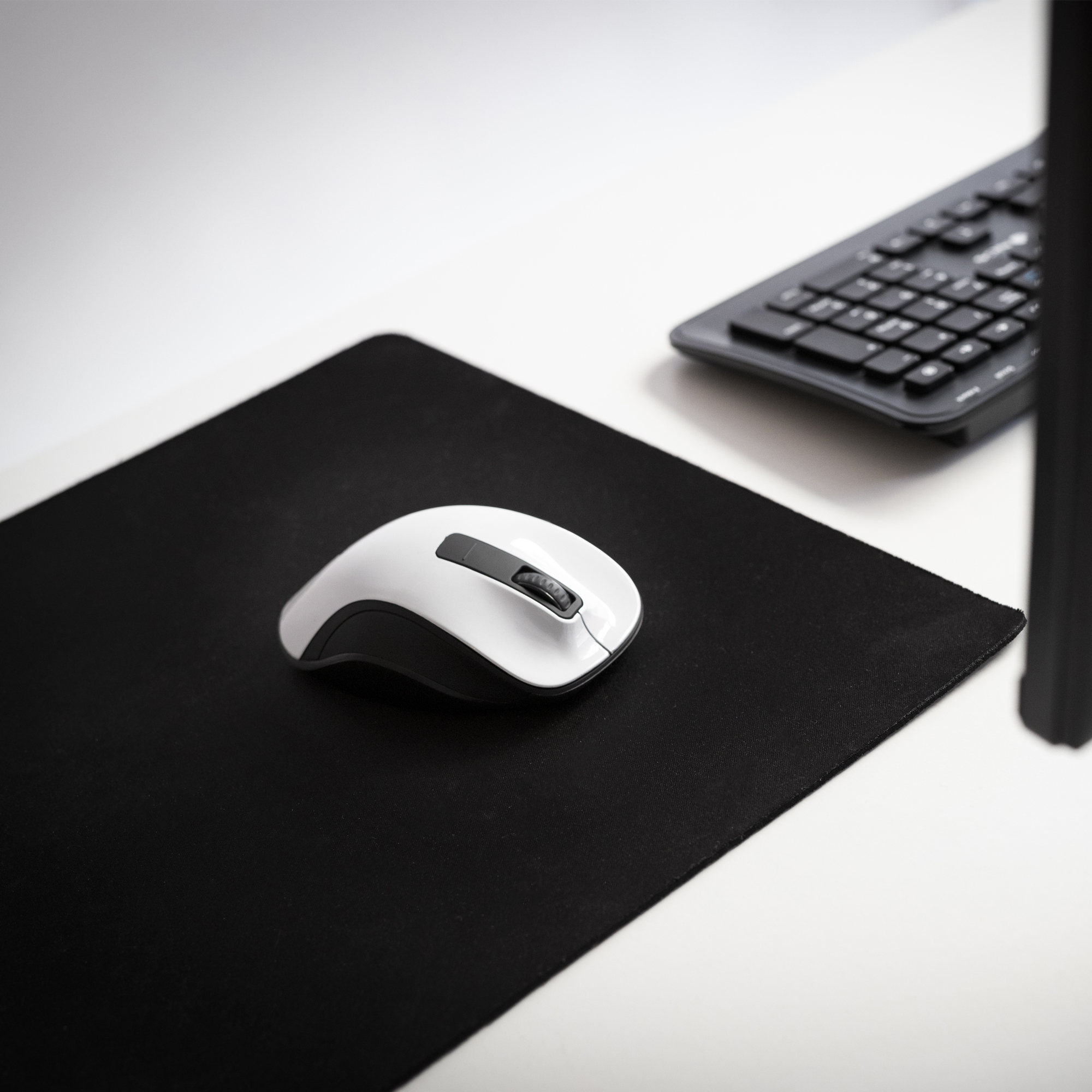 Mouse bianco su tappetino nero, con tastiera sullo sfondo, su scrivania bianca.