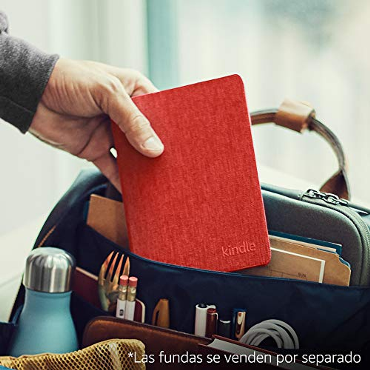 Una mano che tiene un e-reader Kindle rosso in una borsa con oggetti. Sfondo bianco.