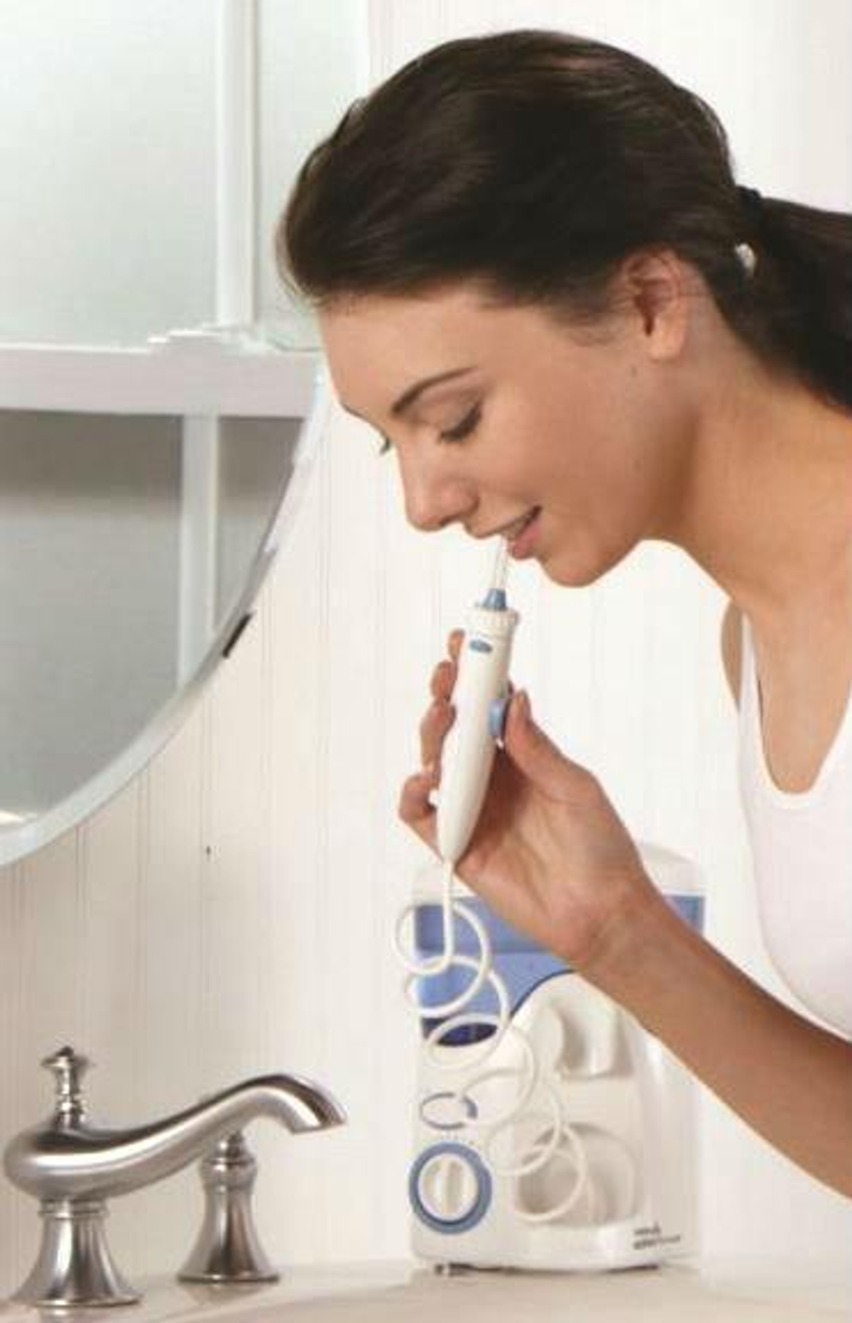 Una mujer usando un irrigador bucal en un baño, con espejo y lavabo visibles.