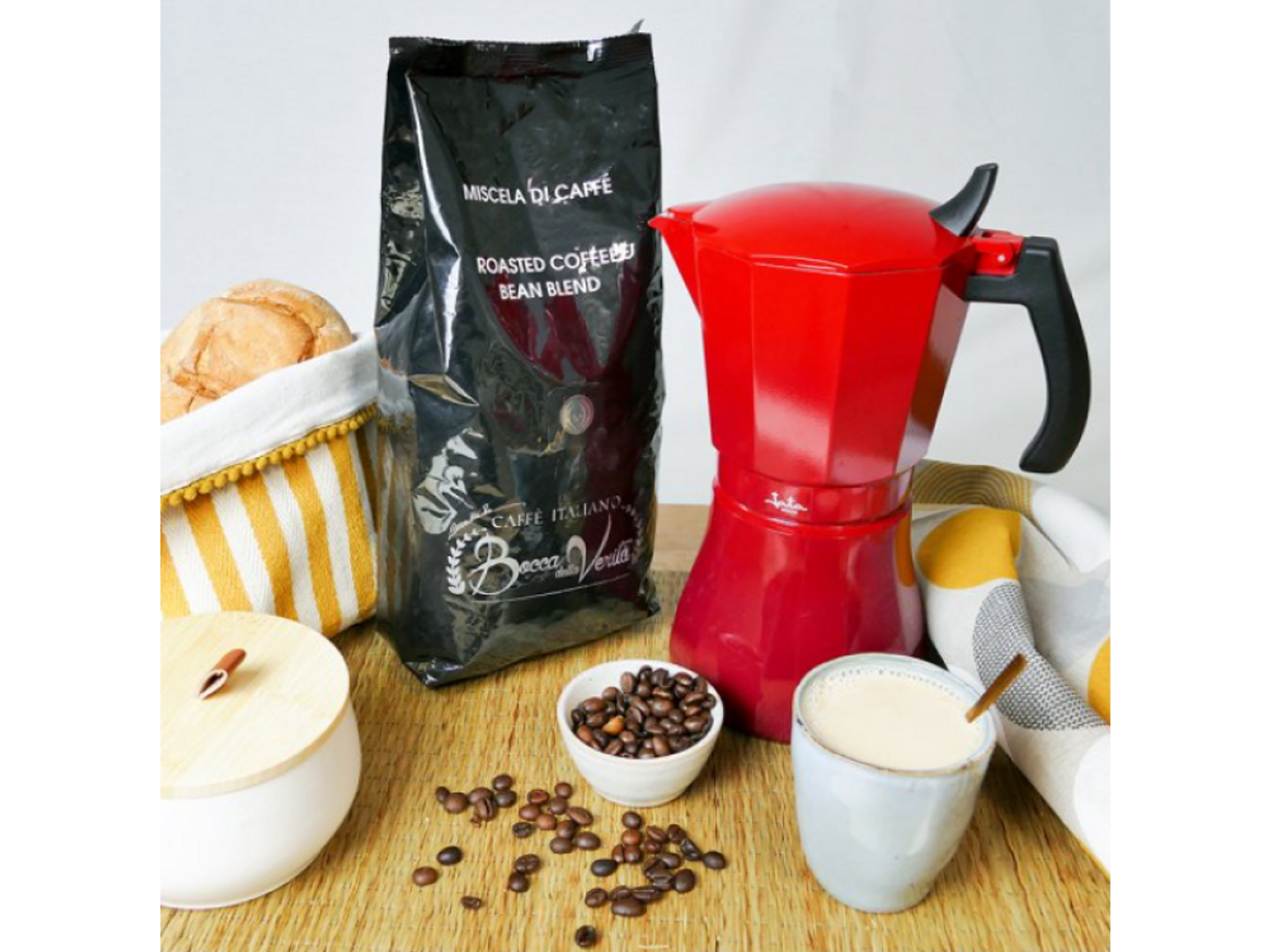 Preparación de café con cafetera roja, granos de café y bolsa. Hay una taza con café y pan.