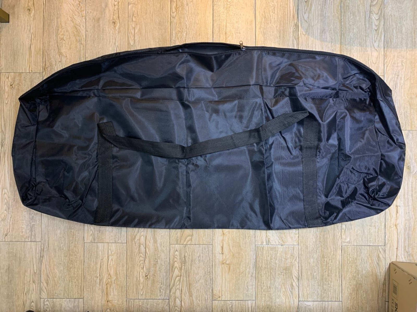 Bolsa de transporte negra y oblonga sobre un suelo con aspecto de madera. Tiene cremallera y correa.