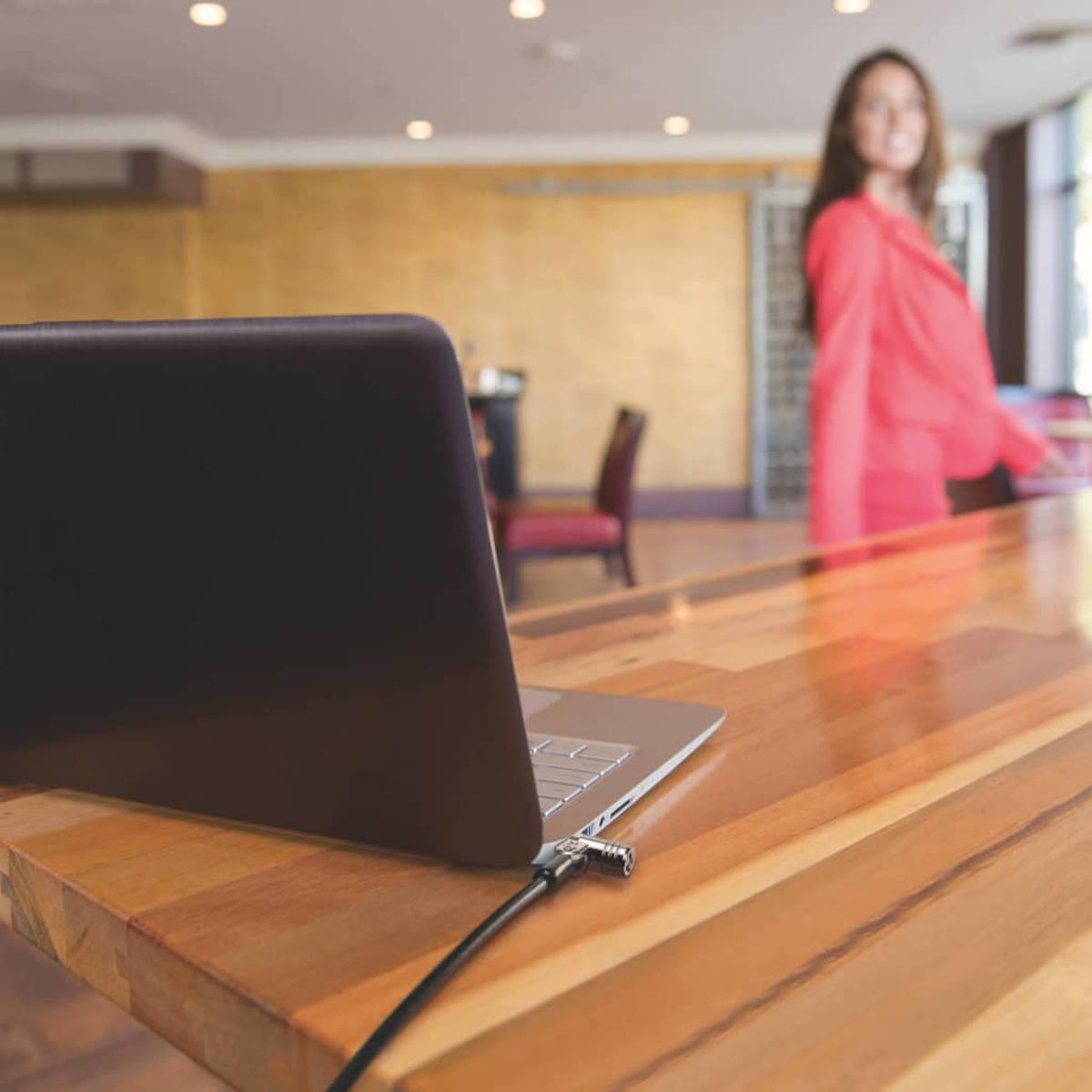 Laptop mit Kabelschloss auf einem Holztisch. Verschwommene Frau im Hintergrund.