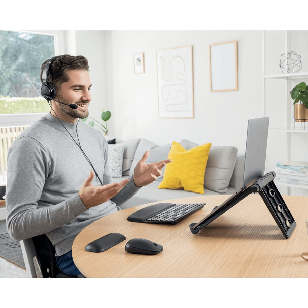 Mann mit Headset, benutzt Laptop auf einem Ständer, lächelt und gestikuliert mit den Händen.