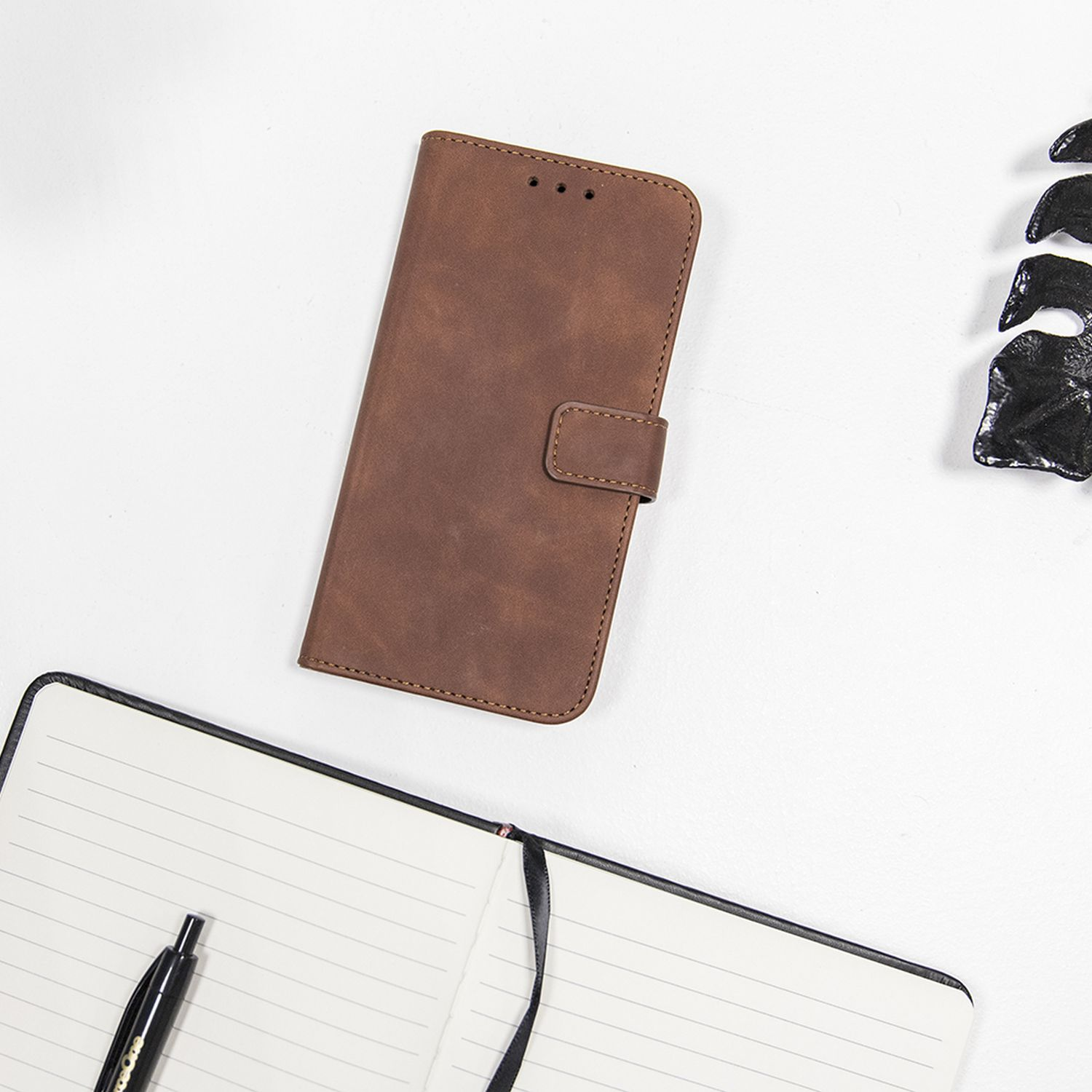 Custodia per telefono in pelle marrone con chiusura magnetica, accanto a un taccuino e una penna neri.