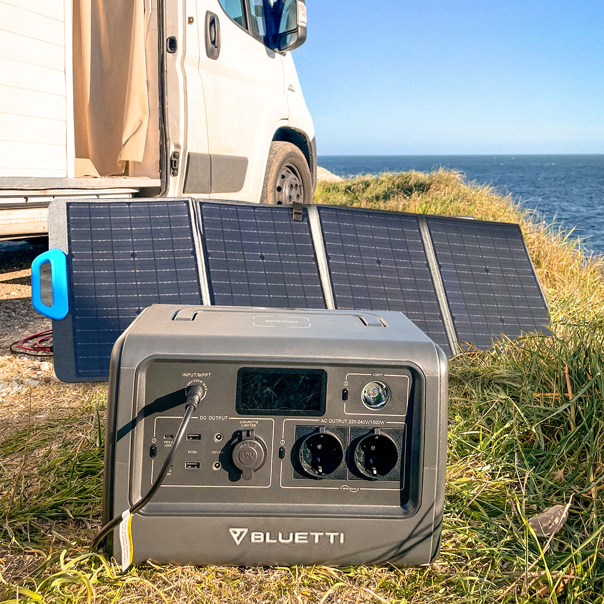 Eine tragbare Stromstation und Sonnenkollektoren liegen auf Gras, in der Nähe eines Wohnwagens und des Meeres.
