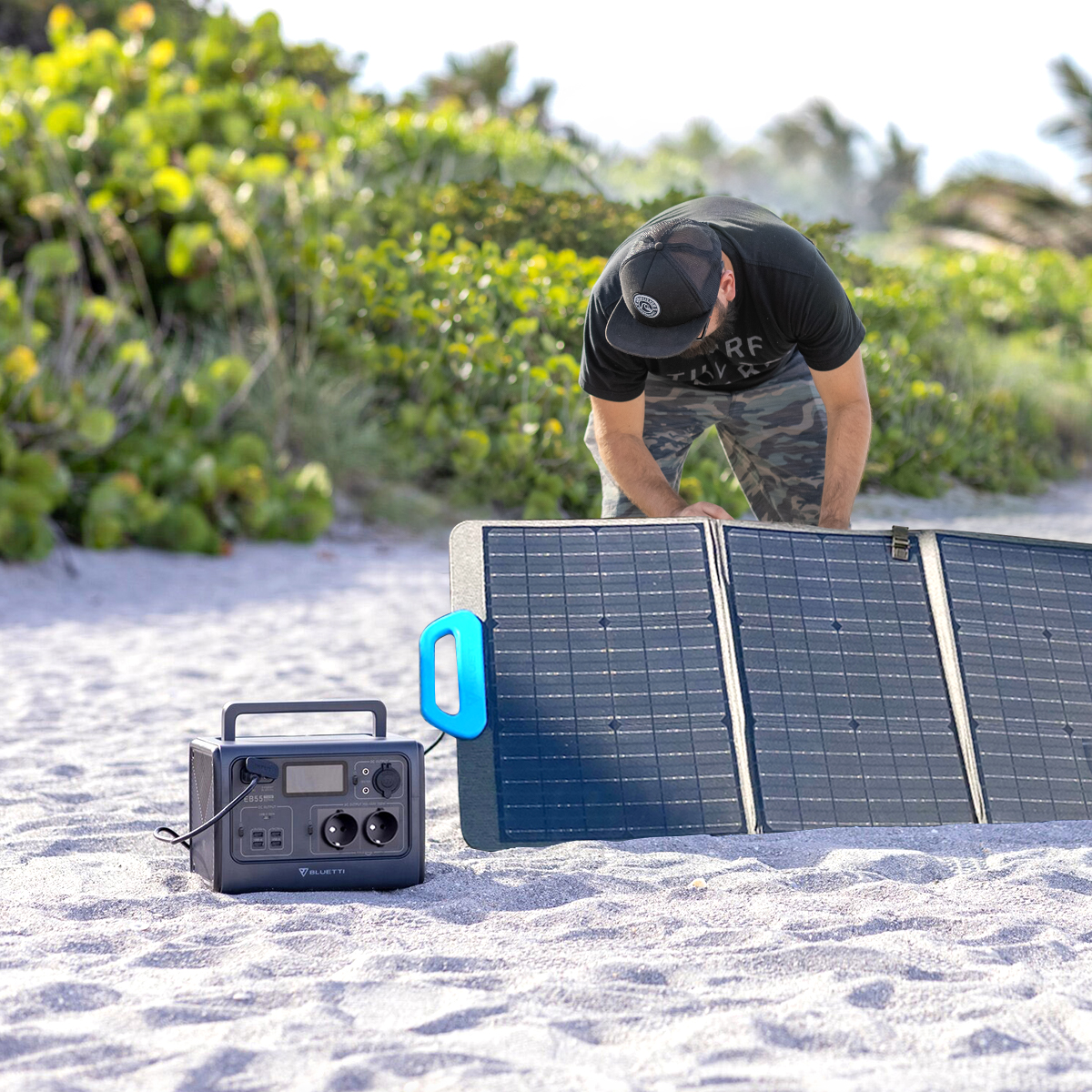 Ein Mann baut ein Solarpanel an einem Sandstrand auf. Ein Generator steht daneben mit blauem Griff.