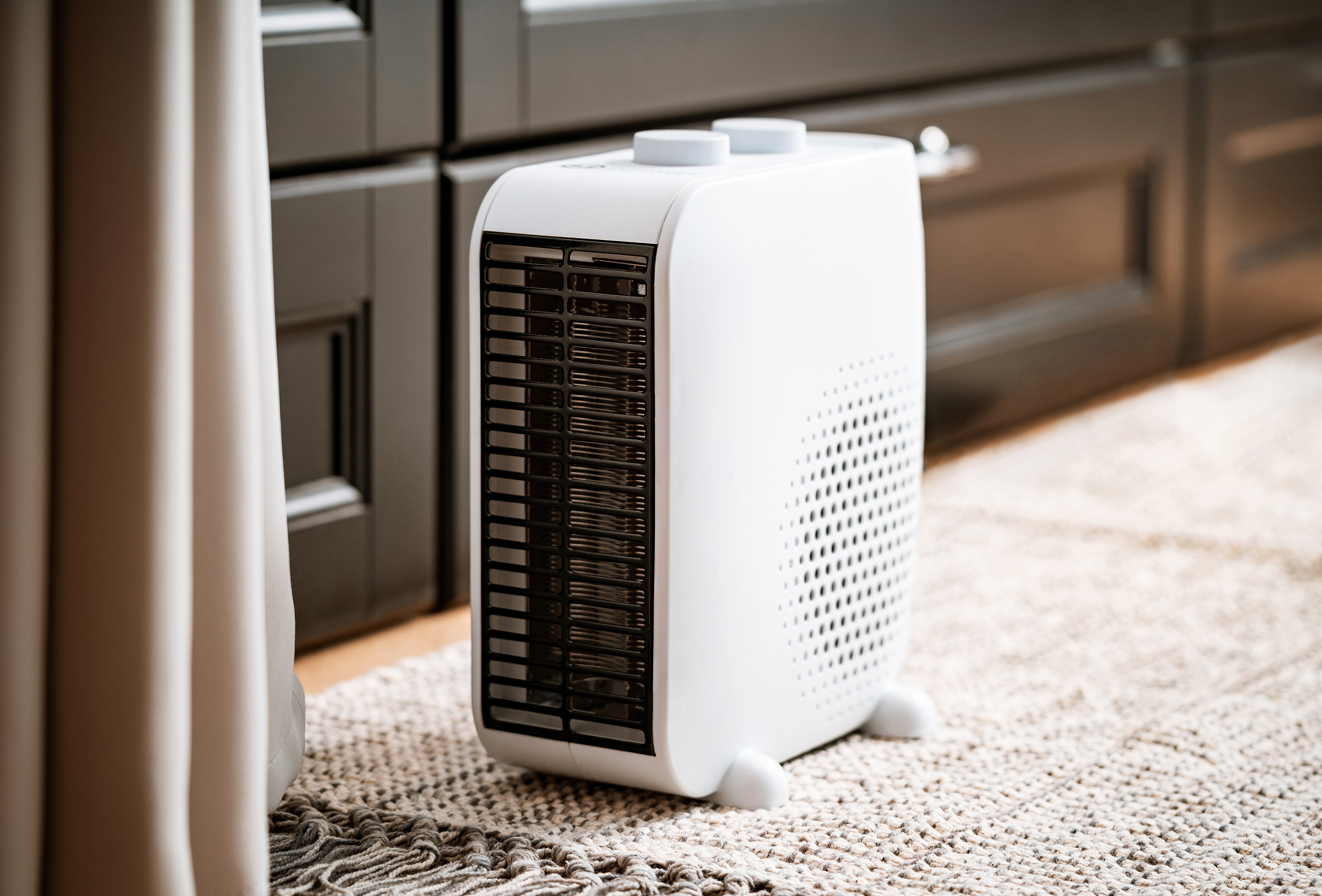 Biały grzejnik z czarną kratką i dwoma pokrętłami. Stoi na dywanie. Ciemne tło.