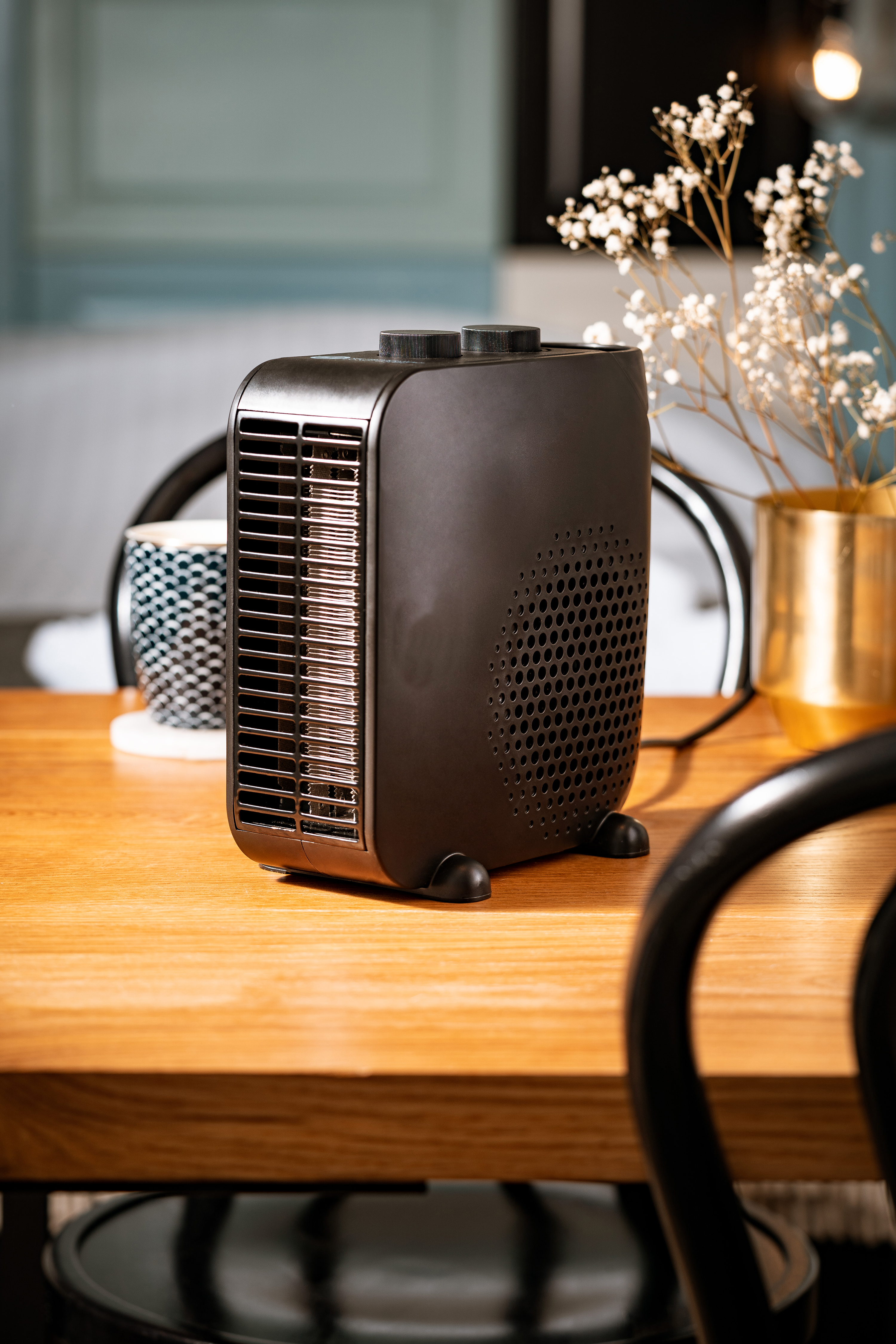 Czarny grzejnik na drewnianym stole. Rozmyte tło z krzesłami, filiżanką i kwiatami.