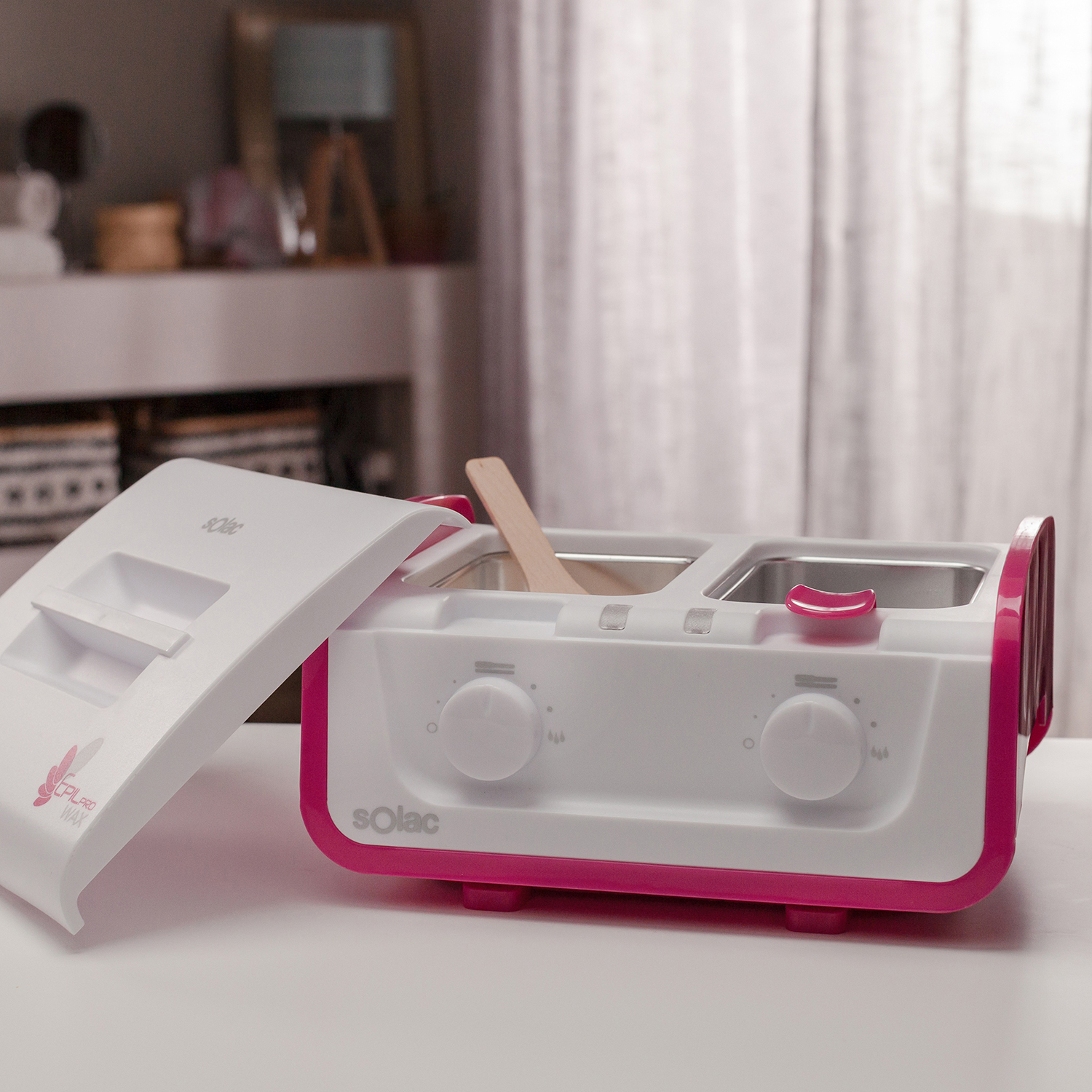 Máquina de cera depilatoria blanca y rosa, tapa abierta, con palito de madera. El fondo son cortinas blancas y crema.