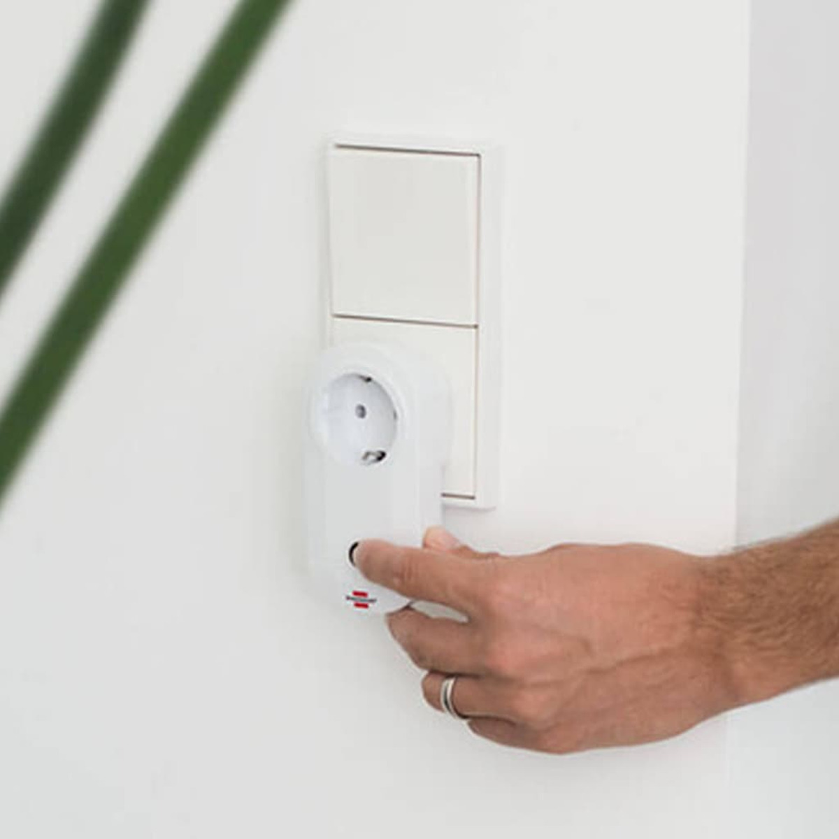 Eine Hand steckt einen weißen Netzadapter in eine Steckdose. Eine weiße Wand und ein Lichtschalter sind sichtbar.